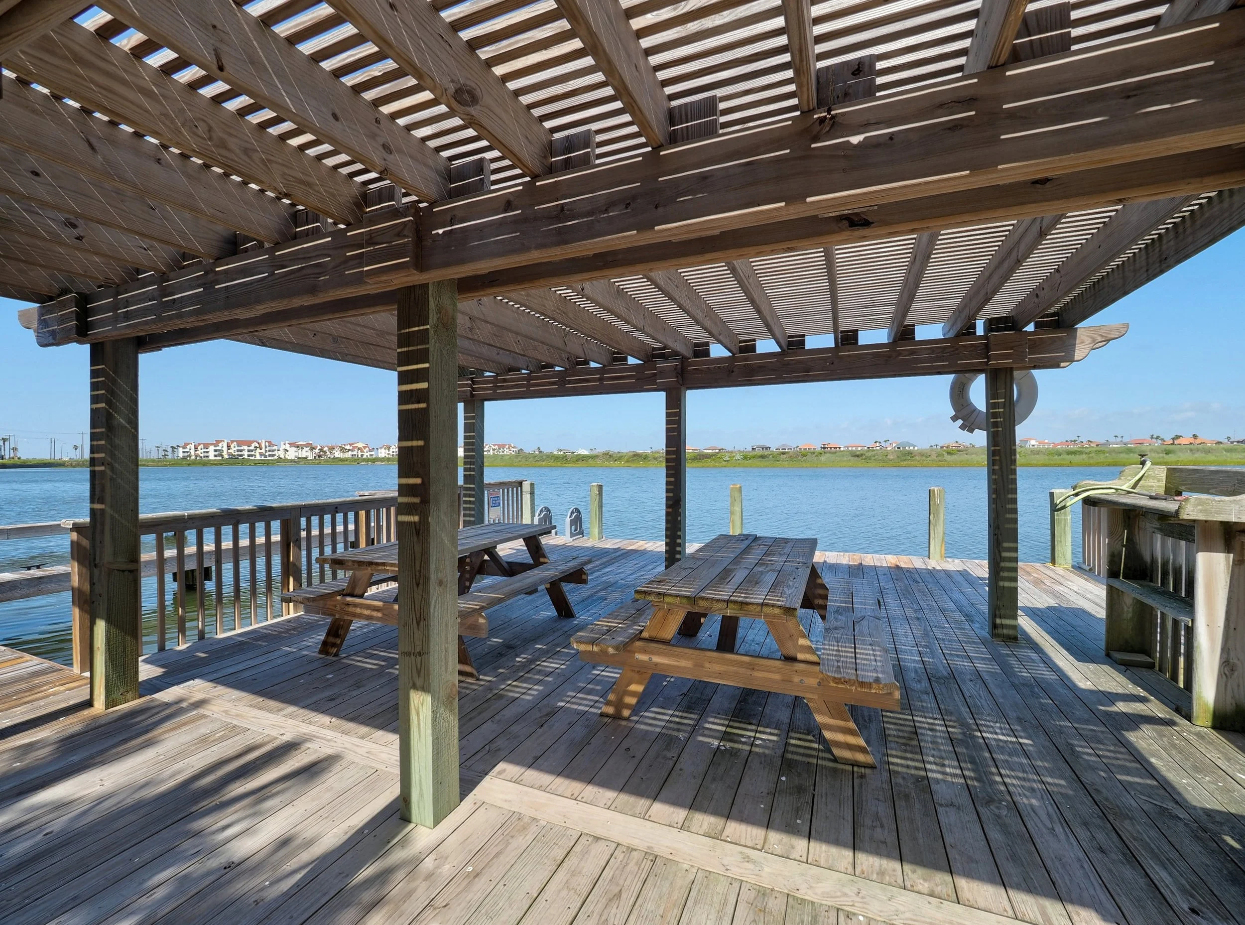 A wooden dock with picnic tables and benches, covered by a wooden roof, overlooking a body of water with houses and buildings in the distance under a clear blue sky.