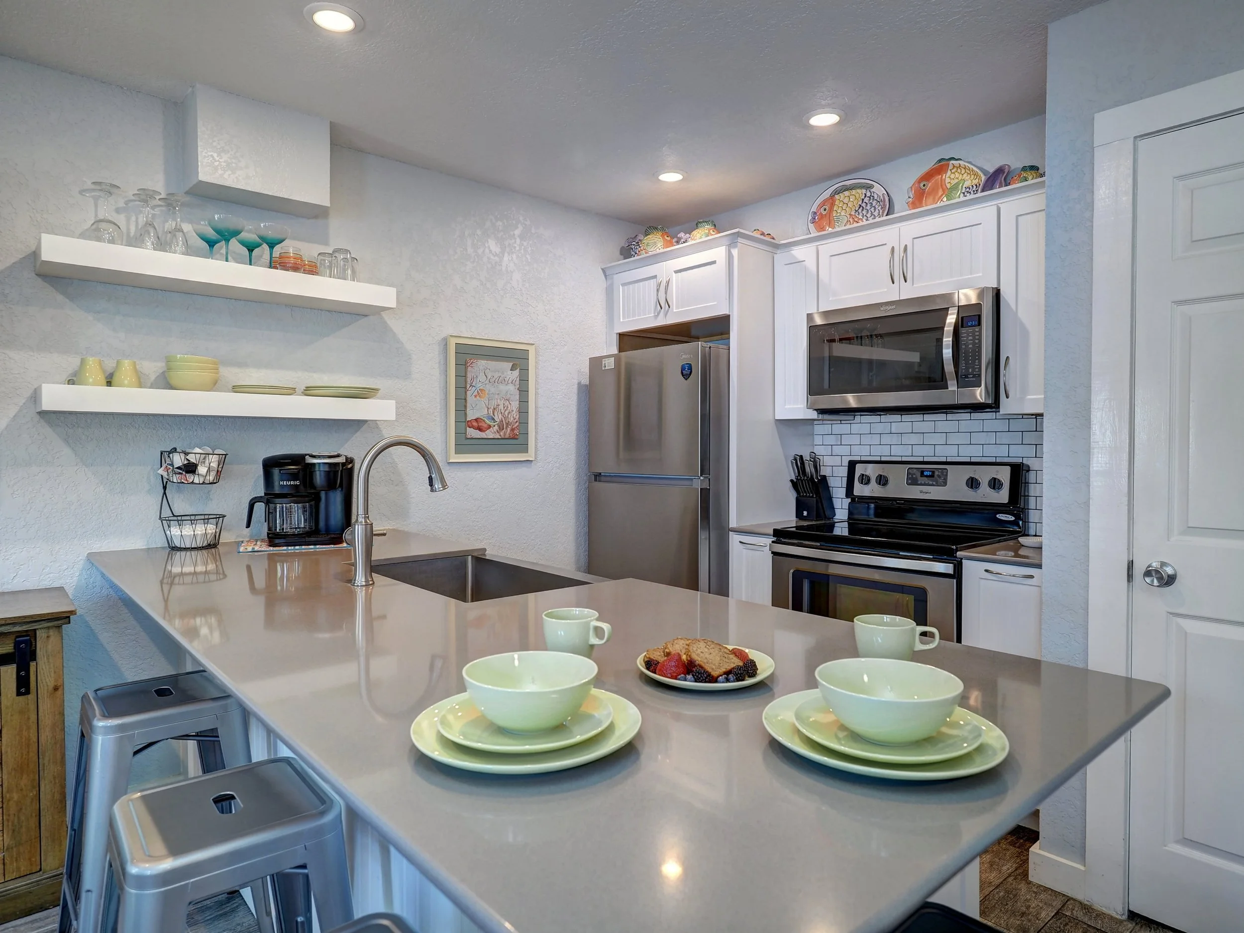 Kitchen with white cabinets, stainless steel microwave and refrigerator, open shelving with colorful fish plates, black electric stove, and breakfast bar with four lime green bowls, cups, and a plate of toast with berries. Recessed lighting and frame