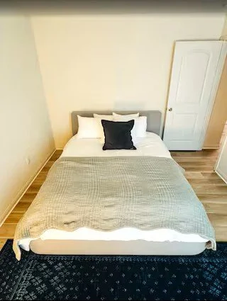 Bedroom with a bed covered by a gray blanket, two white pillows, a black decorative pillow, and a white wardrobe in the corner.