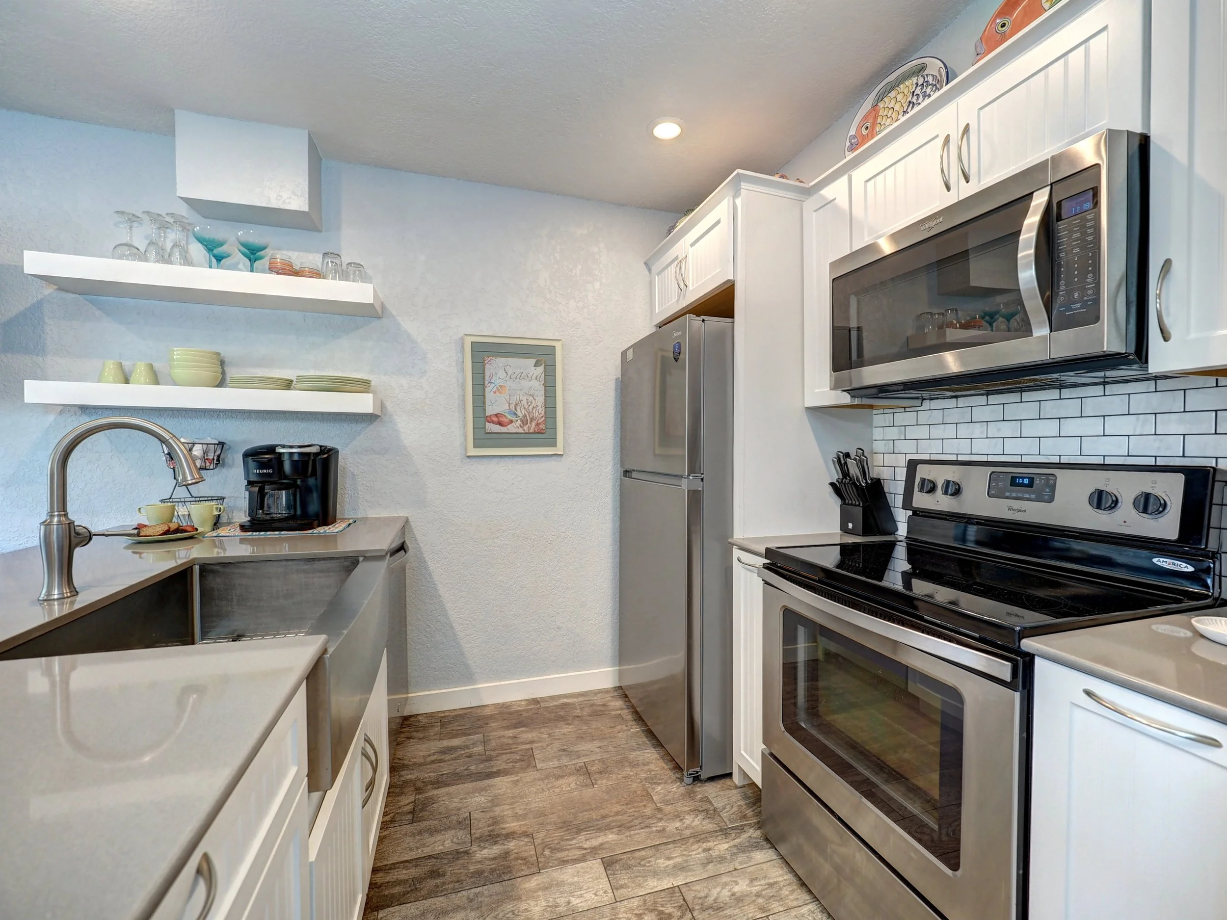 A modern kitchen with white cabinetry, a stainless steel microwave above a stove, open shelves with dishes, and a black coffee maker on the counter. There is a framed ocean-themed picture on the wall.