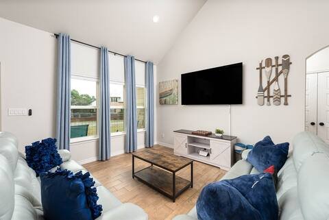 Living room with white walls and ceiling, large window with blue curtains, wall-mounted flat screen TV, rustic wall decor, white entertainment unit, wooden coffee table, white couches with blue and dark throw pillows.