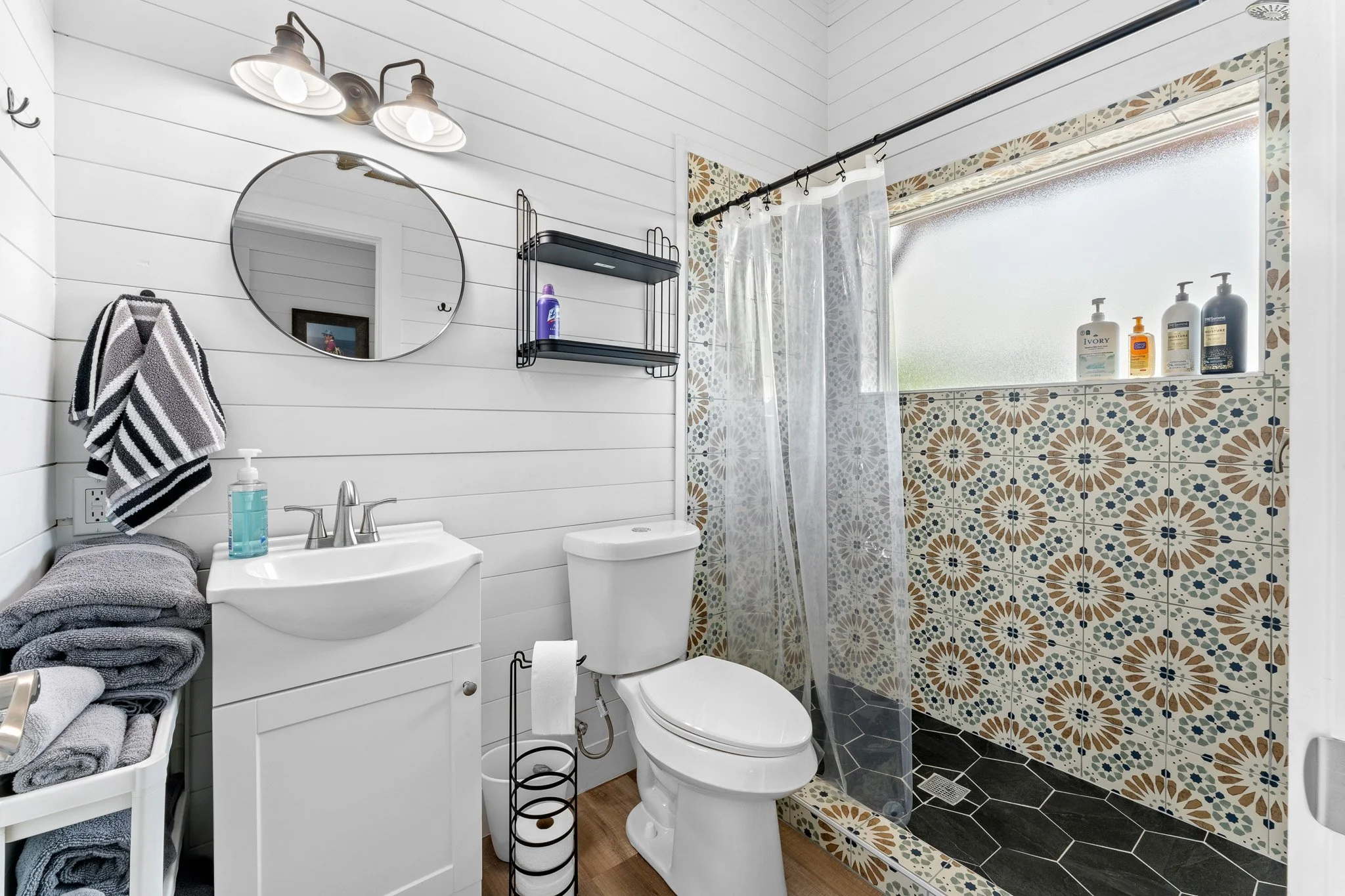 A small bathroom with white-paneled walls, a round mirror, black metal shelves, a white sink, a toilet, and a shower area with patterned tiles and a clear glass door. Shelves hold toiletries, and towels are stacked on a laundry basket.