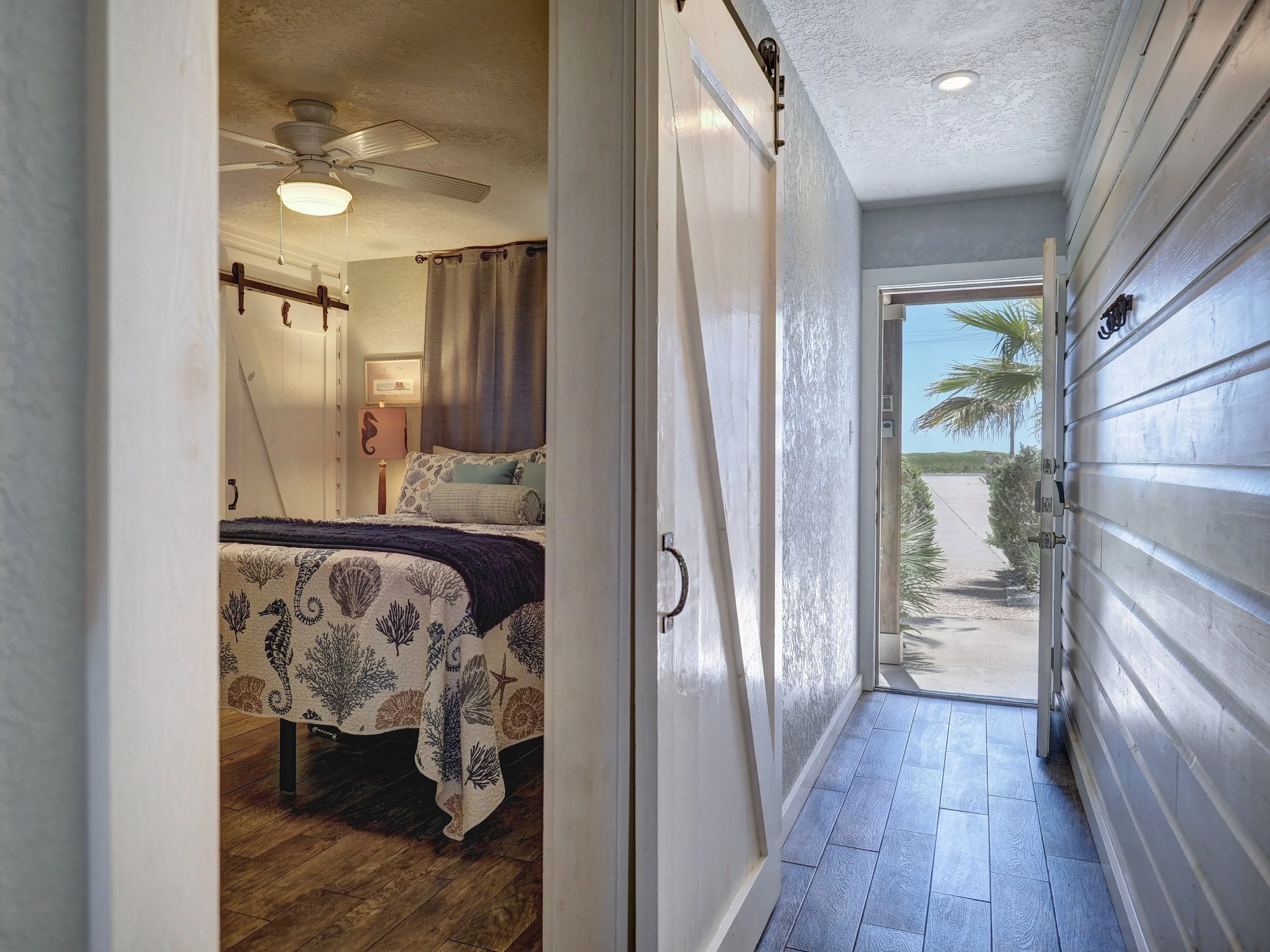 View of a bright, sunny porch with a palm tree visible outside through an open door, with a glimpse of ocean or lake neighboring the landscape.