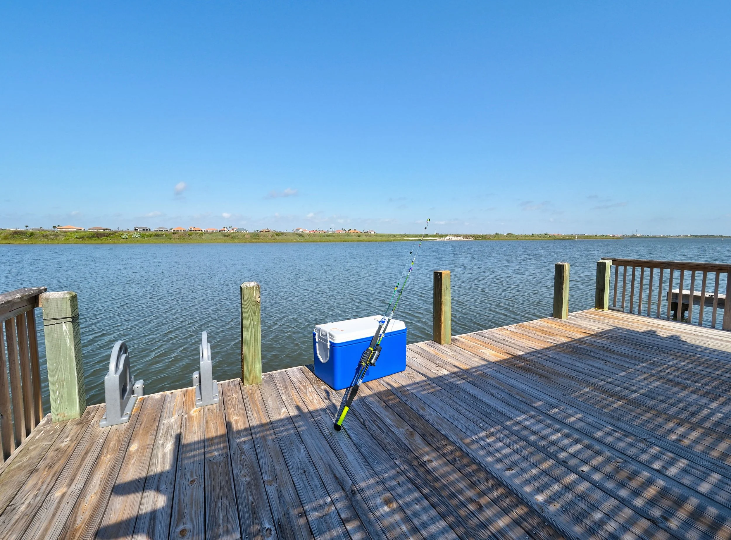 A view from a wooden dock over a body of water, with a blue cooler, fishing rod, and chairs in the foreground. Houses are visible on the opposite shoreline under a clear blue sky.