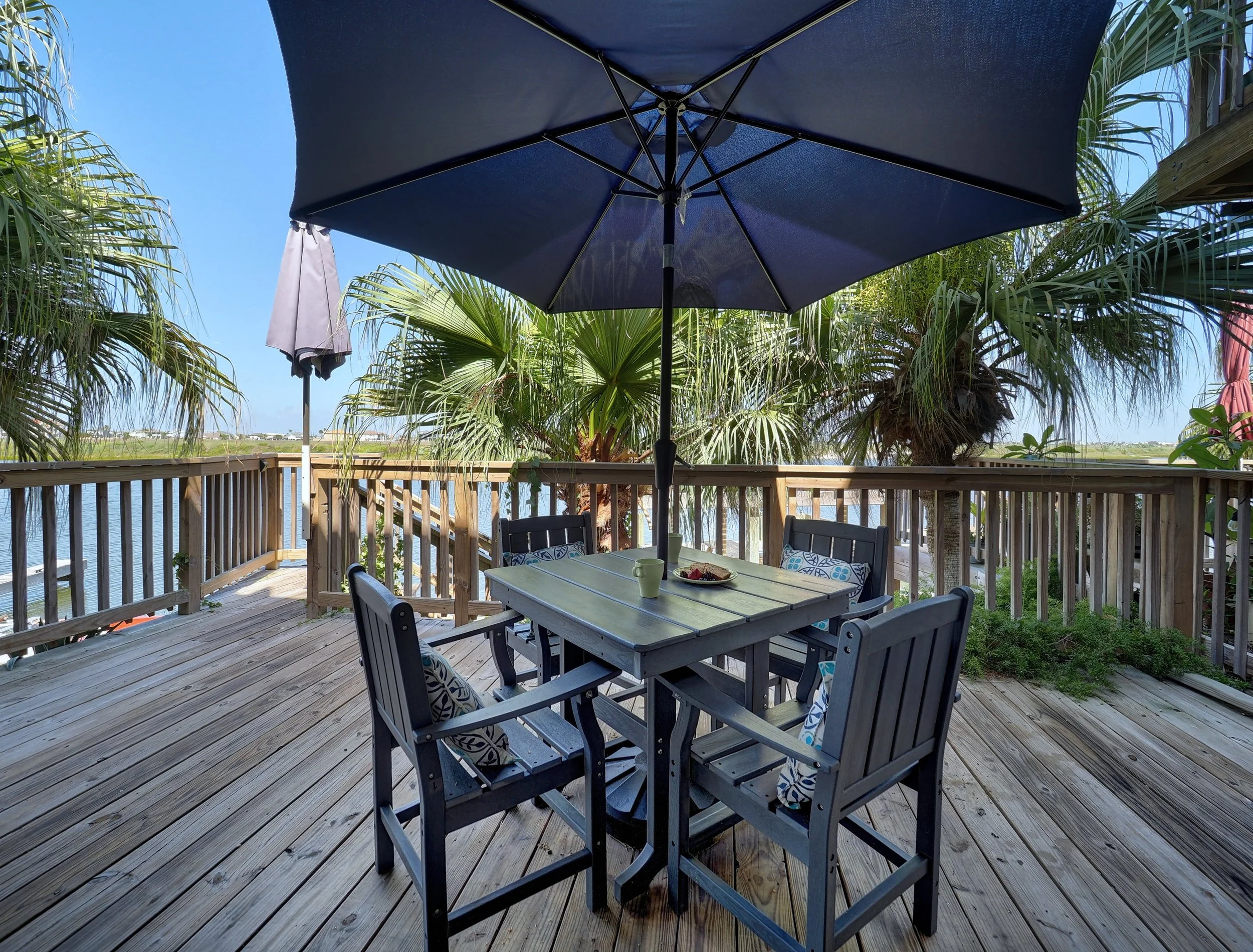 A wooden deck with a black dining table, chairs, and a large blue patio umbrella outdoors, surrounded by palm trees and overlooking a body of water under a clear blue sky.