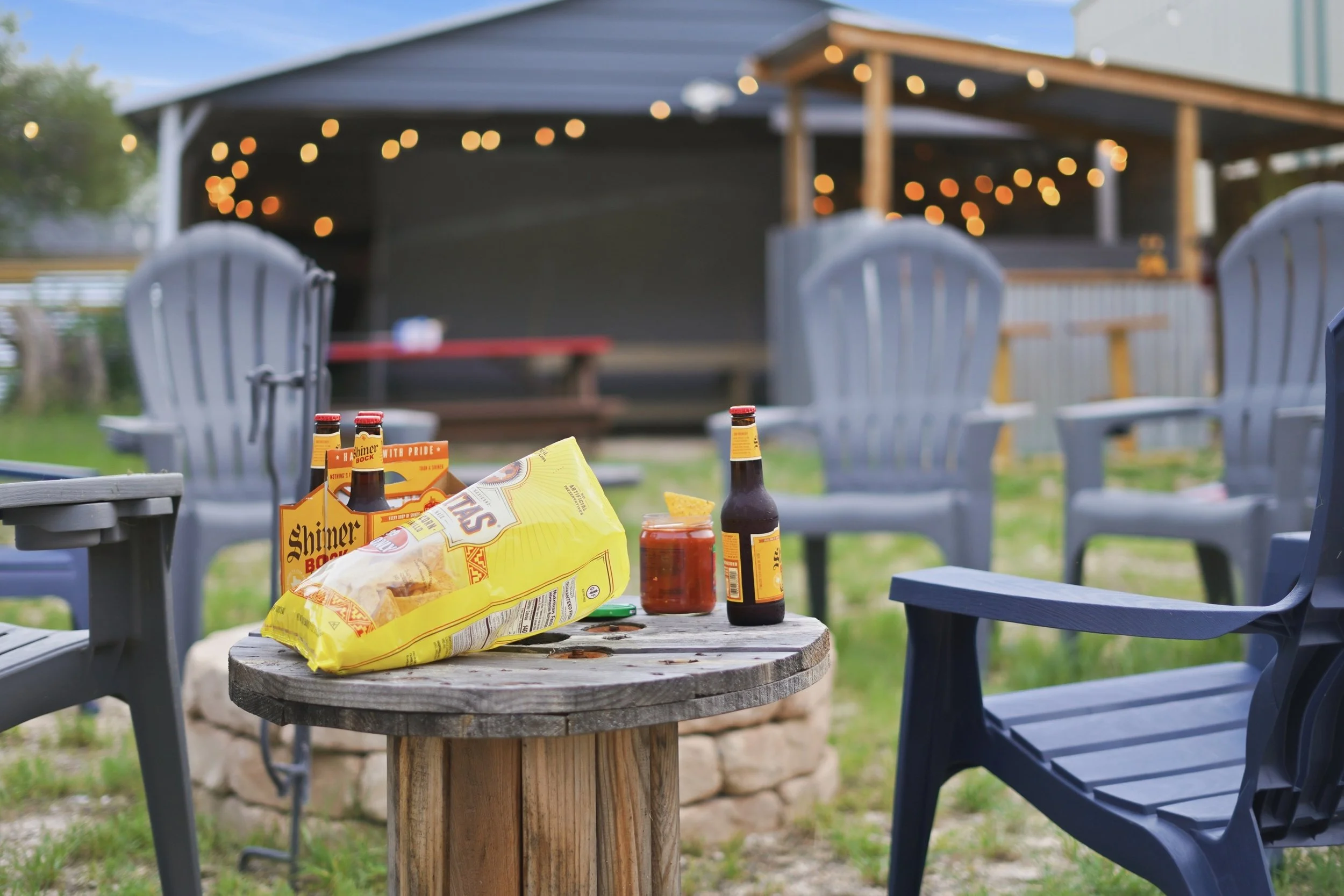 Outdoor patio setup with a wooden table holding snacks and drinks, surrounded by Adirondack chairs, with a covered pavilion in the background decorated with string lights.