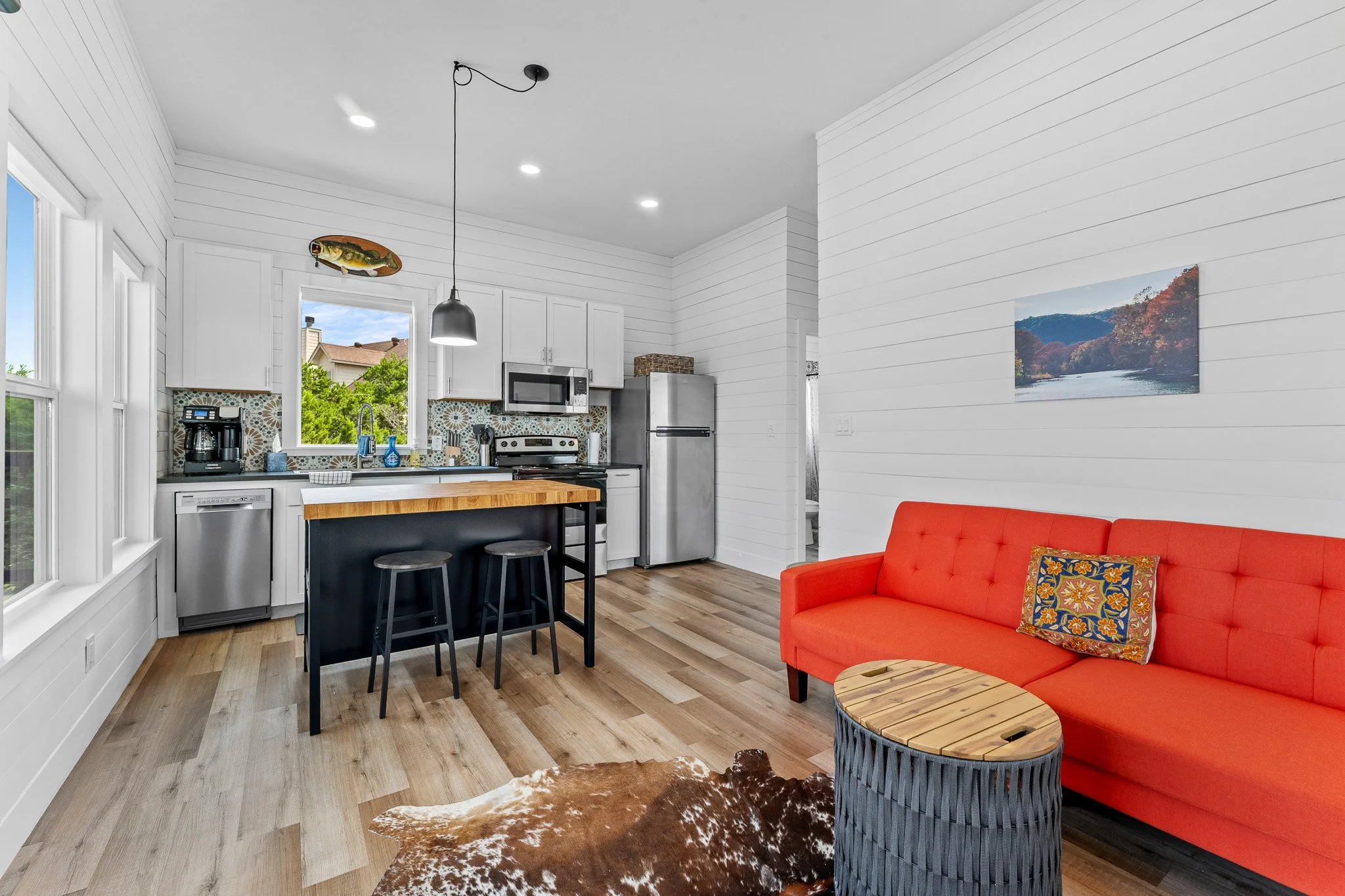 Open-concept living room and kitchen with white walls and cabinets, wooden flooring, and a red sofa with a colorful pillow.