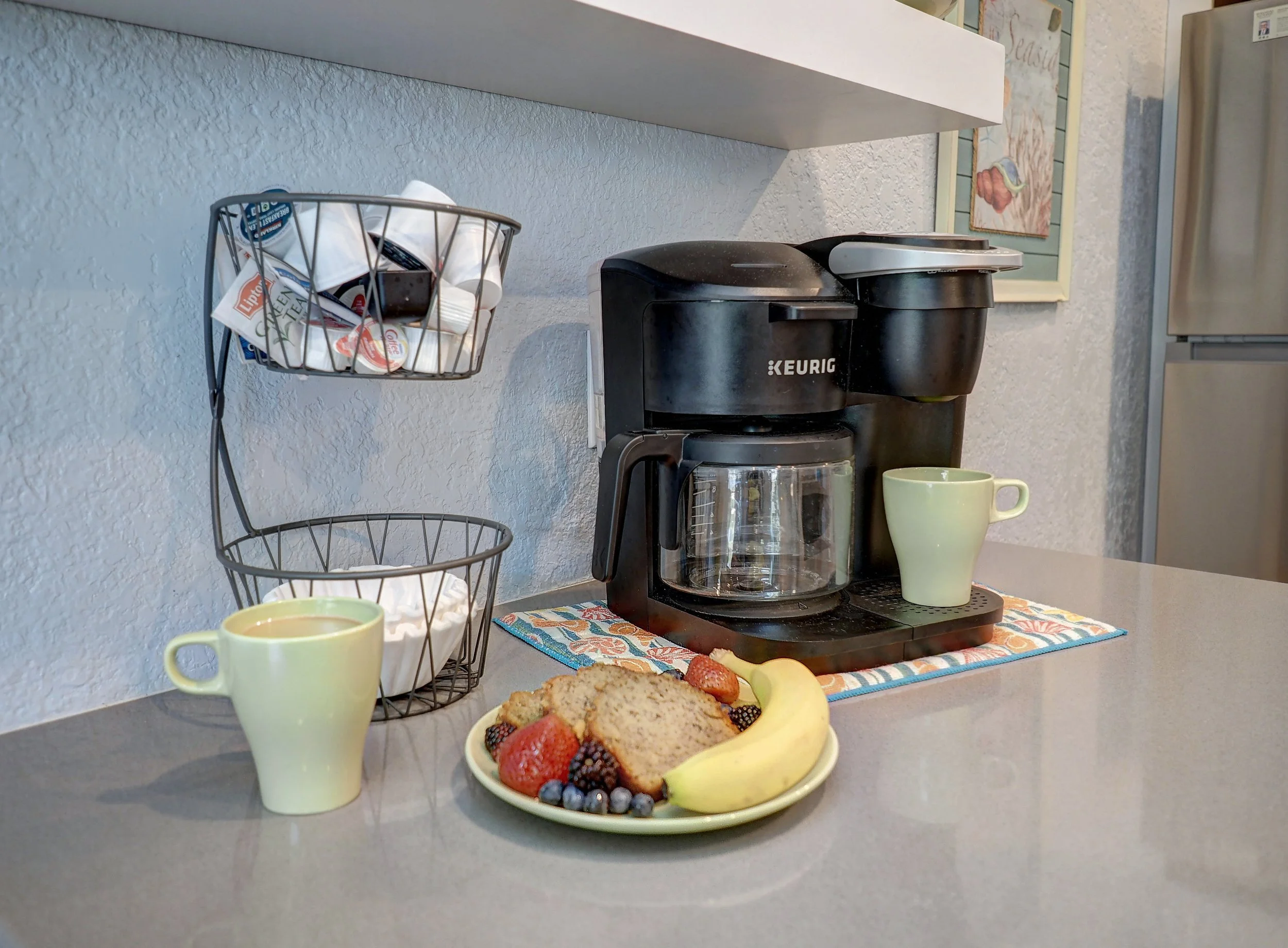 Breakfast setup with a coffee maker, two yellow mugs, a plate of cookies and fresh fruit, and a wire basket with various small containers, all placed on a kitchen counter with a colorful towel underneath.