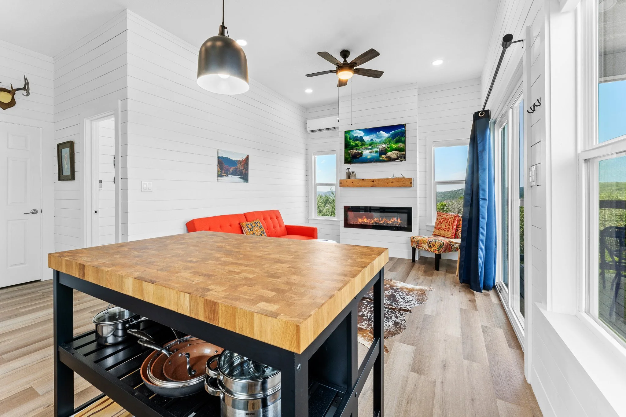 Living room with white shiplap walls, a ceiling fan, wall-mounted tv, fireplace, large window with blue curtains, and a wooden kitchen island with a cookware rack underneath.