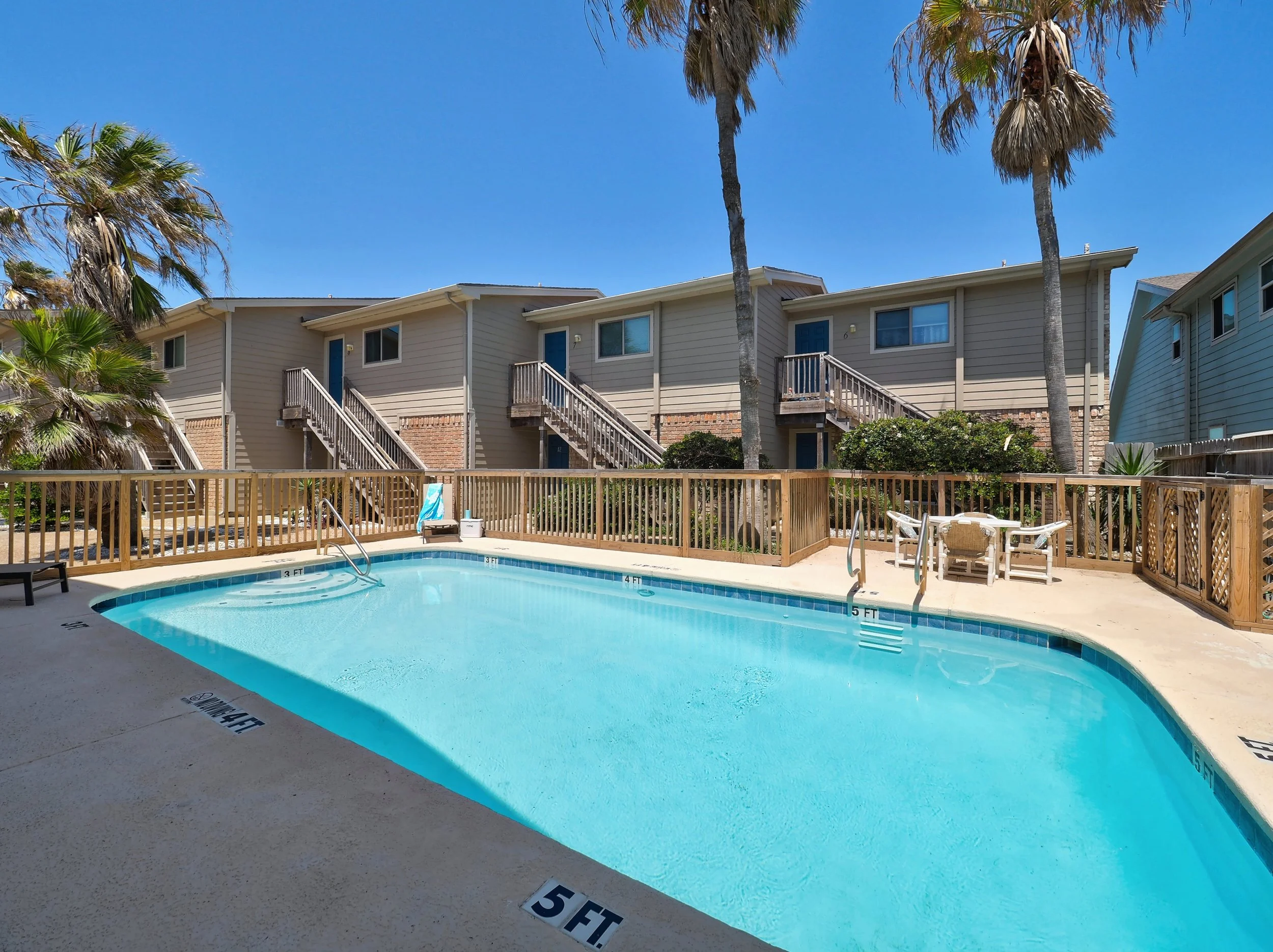 A swimming pool with clear blue water surrounded by a wooden fence, with two-story houses and palm trees in the background under a bright blue sky.
