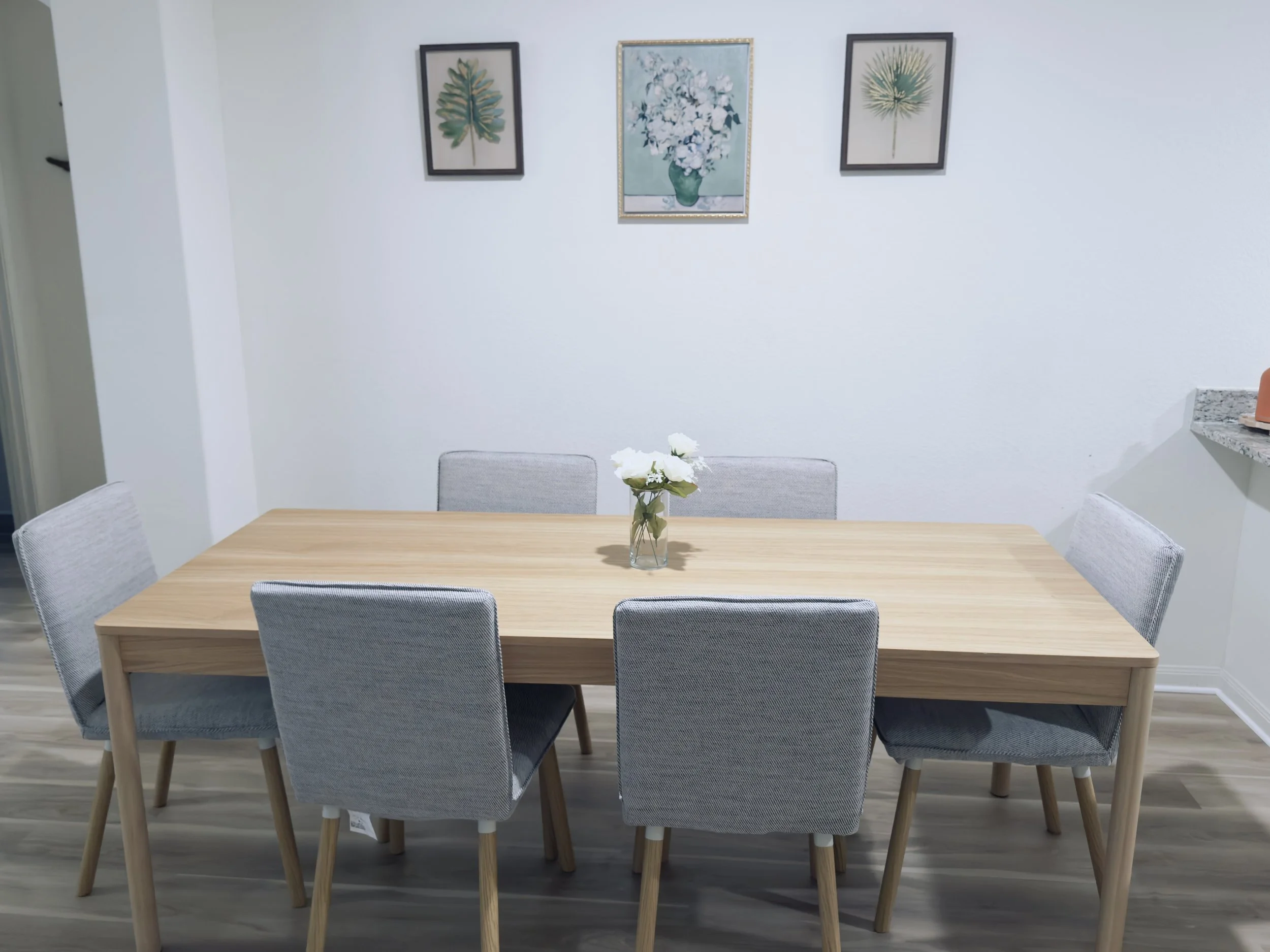 A dining room with a light wooden table, surrounded by six gray upholstered chairs with wooden legs. On the table, there is a glass vase with white flowers. The background features a white wall with three framed botanical prints and a central painting of white flowers in a teal vase.