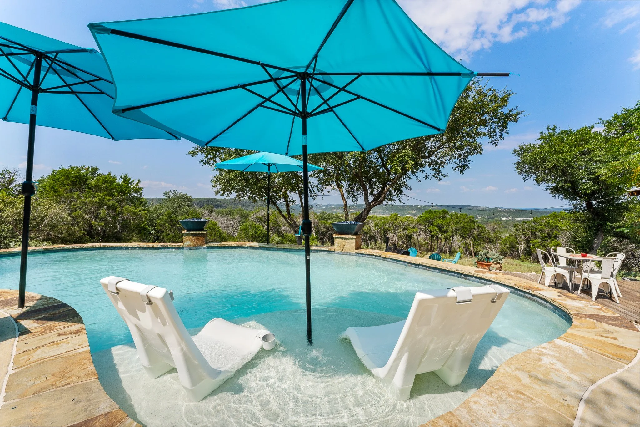Swimming pool with two white lounge chairs in the foreground and two blue umbrellas providing shade. There are green trees and a distant landscape under a blue sky with some clouds.
