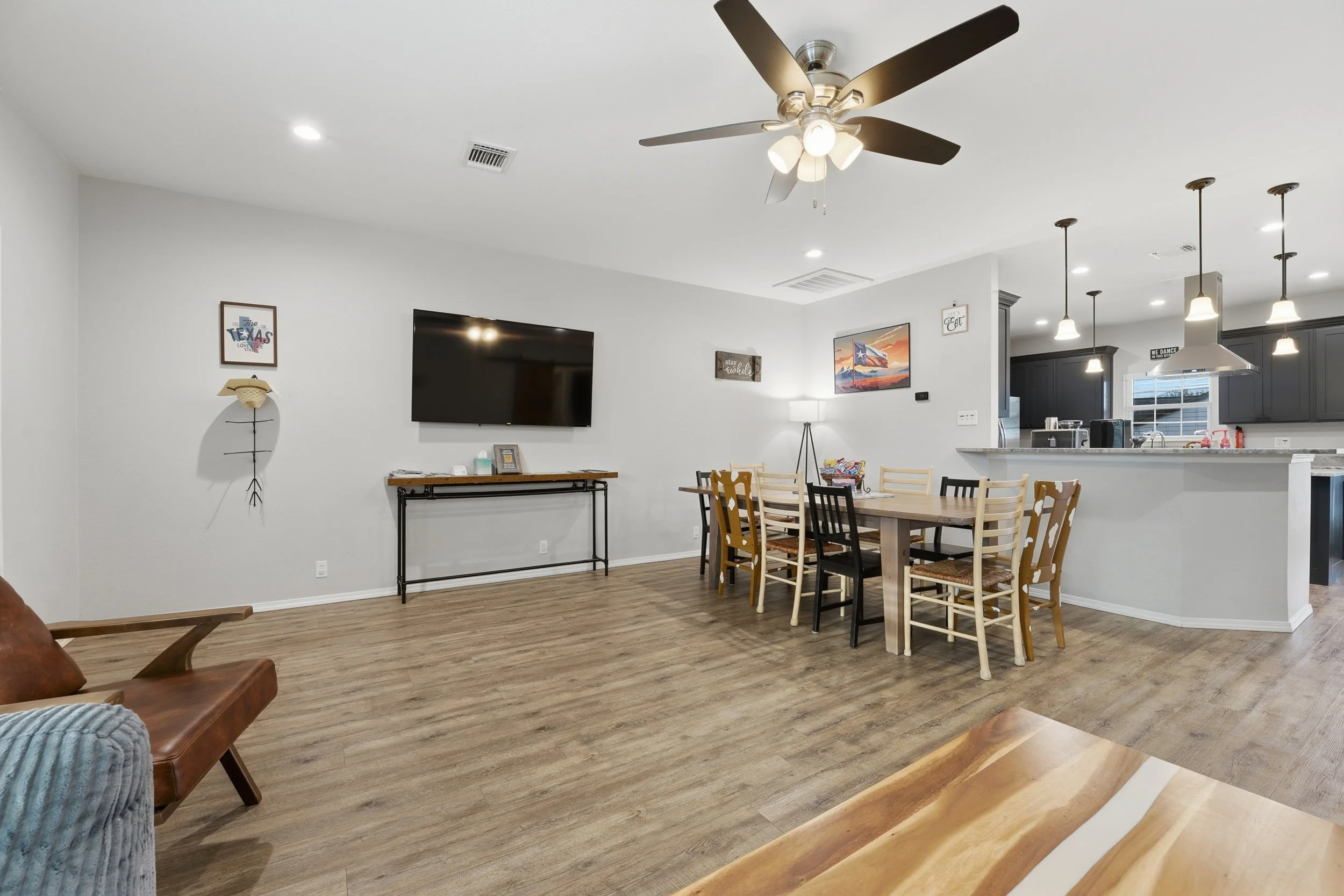 Open-concept living and dining area with a mounted flat-screen TV, framed wall art, a wooden dining table with six chairs, a kitchen with dark cabinets, pendant lights, and large windows.