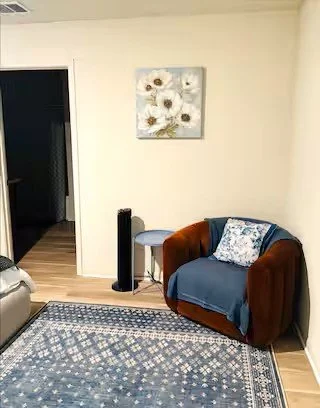 A cozy living room corner with a blue armchair draped with a blue blanket and patterned pillow, a small side table, a floor fan, a framed flower painting on the wall, and a patterned rug on the hardwood floor.