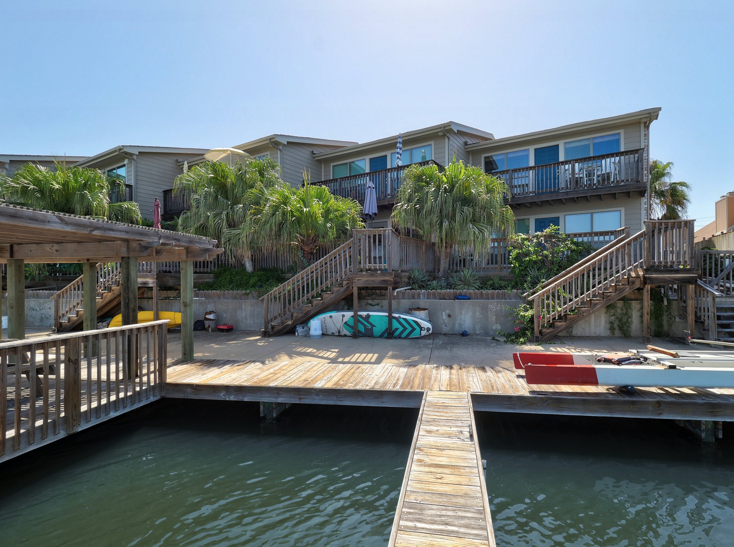 Multi-level wooden deck and stairs with a house and palm trees in the background, next to a waterway with a boat docked underneath.