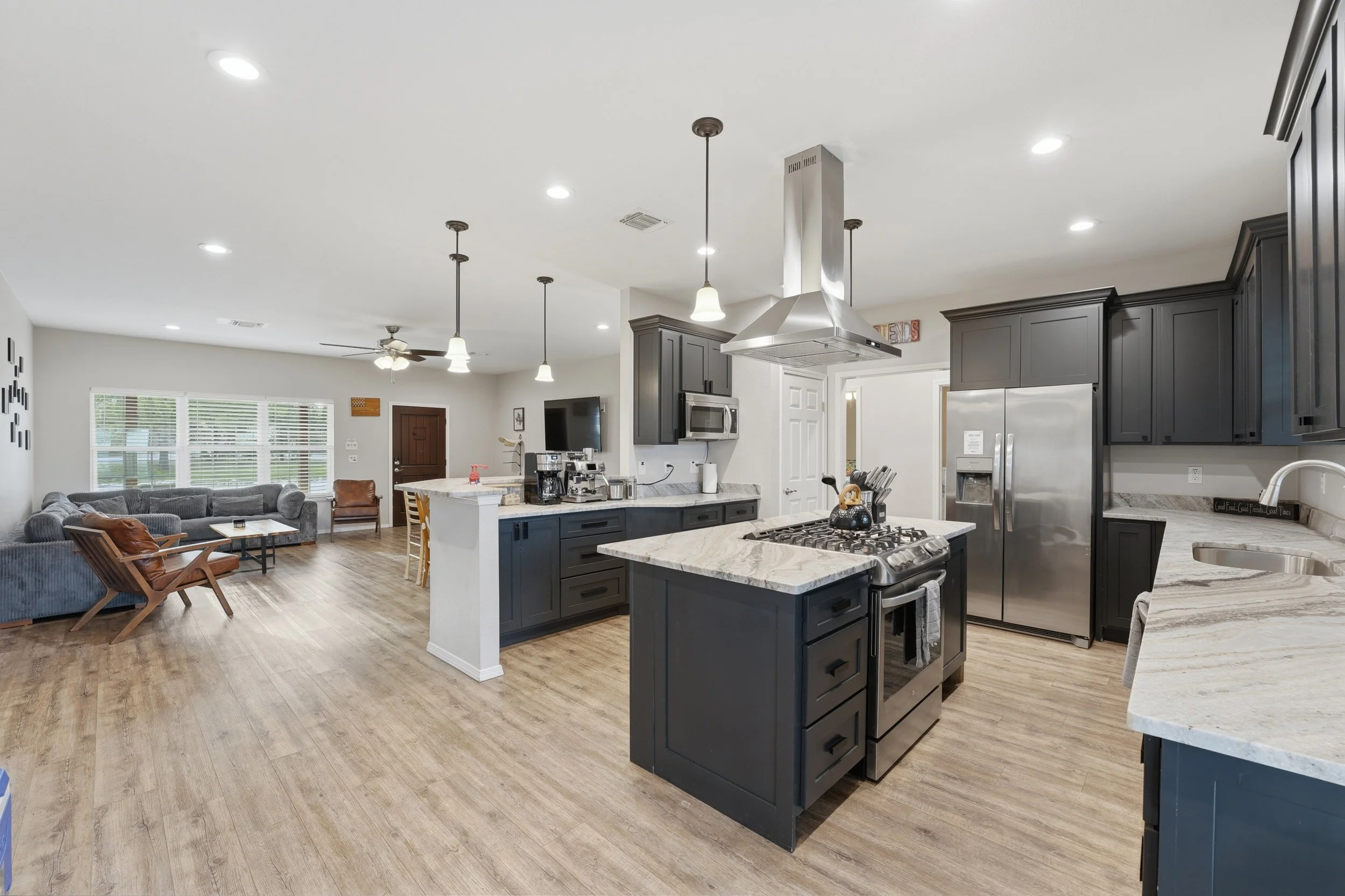 Open-concept kitchen and living room with gray cabinets, stainless steel appliances, a gas stove island, dining chairs, sofa, chairs, television, ceiling fans, and hardwood floors.