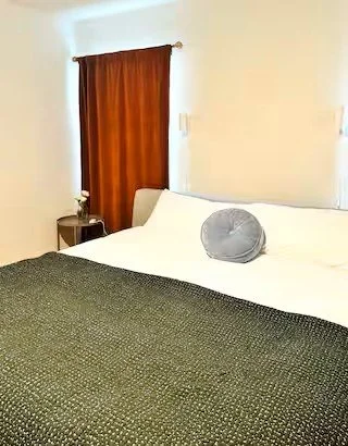 A neatly made bed with white sheets, a gray round pillow, and a dark green coverlet, next to a nightstand with a small white vase and flower, inside a room with a window covered by a red curtain.
