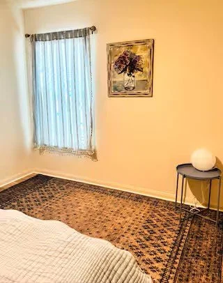 A corner of a bedroom with a window covered by sheer curtains, a painting of a tree on the wall, a side table with a round lamp, and a patterned carpet.