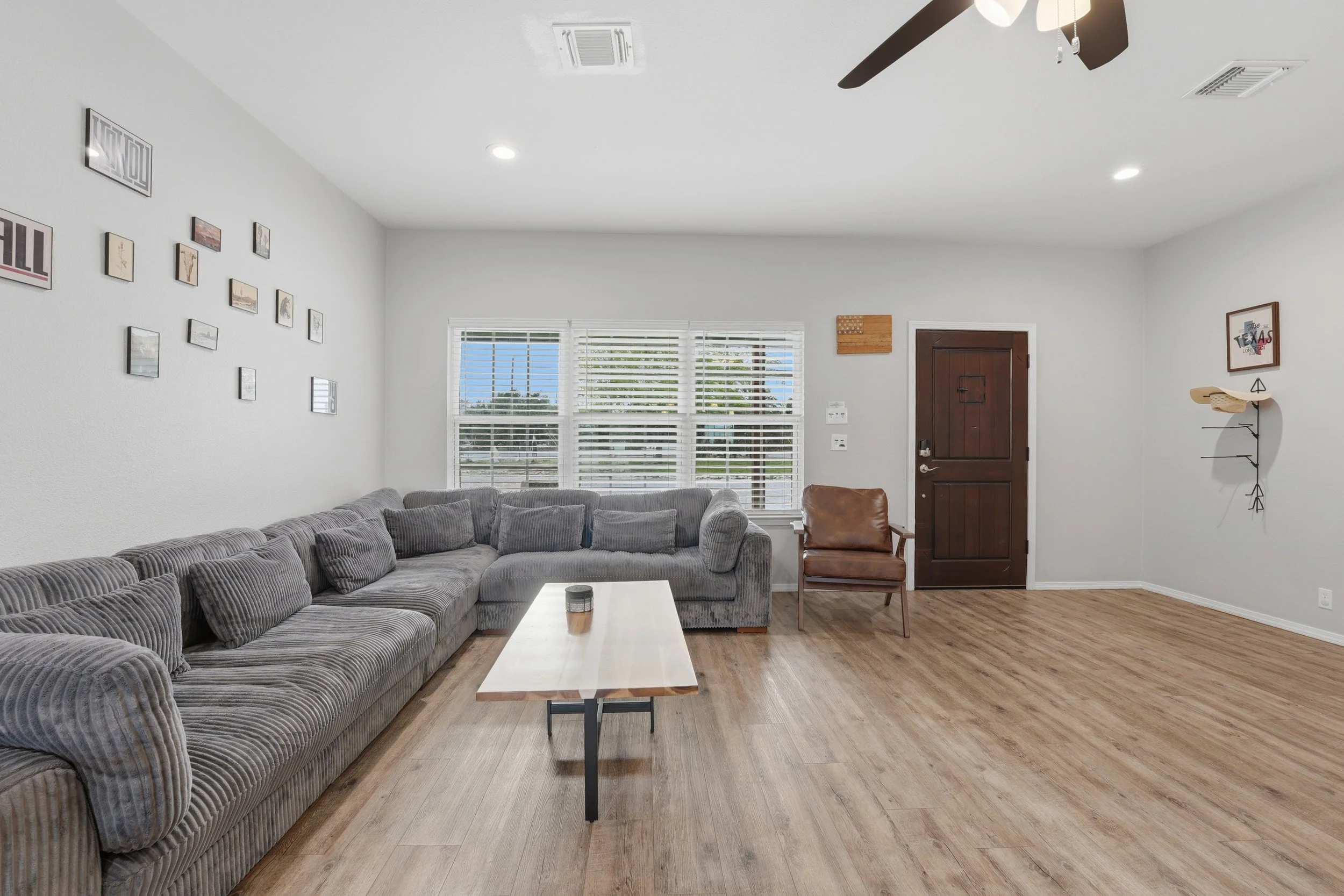 Living room with a large gray sectional sofa, a wooden coffee table, a brown chair, a wall with picture frames, large window with blinds, and a white door with a wooden frame.