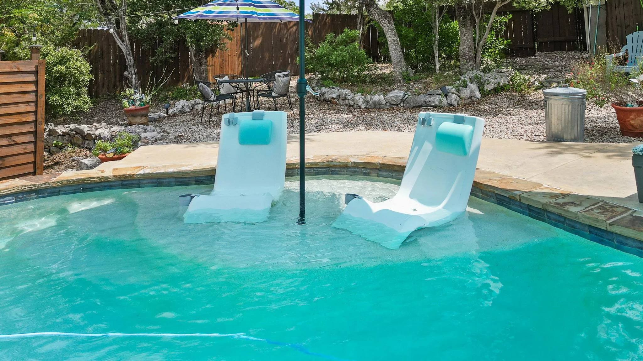 A backyard swimming pool with two white pool floats shaped like lounge chairs in the water. There is a blue and white striped umbrella over the pool, with a small patio table and four chairs nearby. The background features trees, bushes, a wooden fence, and a trash can.