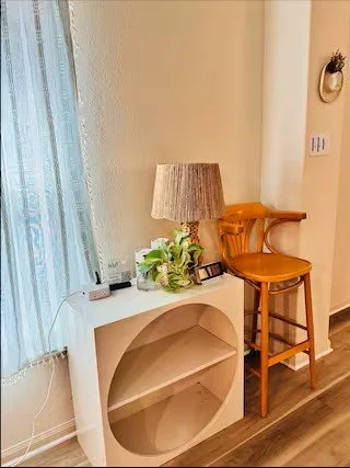 A corner of a room with a white bookshelf, a table lamp with a textured shade, a small plant, and a wooden barstool near a beige wall.