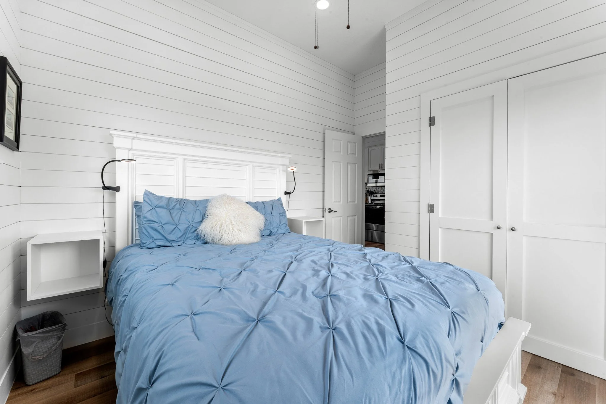A bedroom with white shiplap walls, a white display headboard with a blue tufted comforter, a white furry pillow, and black reading lamps attached to the wall. There are white nightstands on either side of the bed, a doorway leading to another room, 