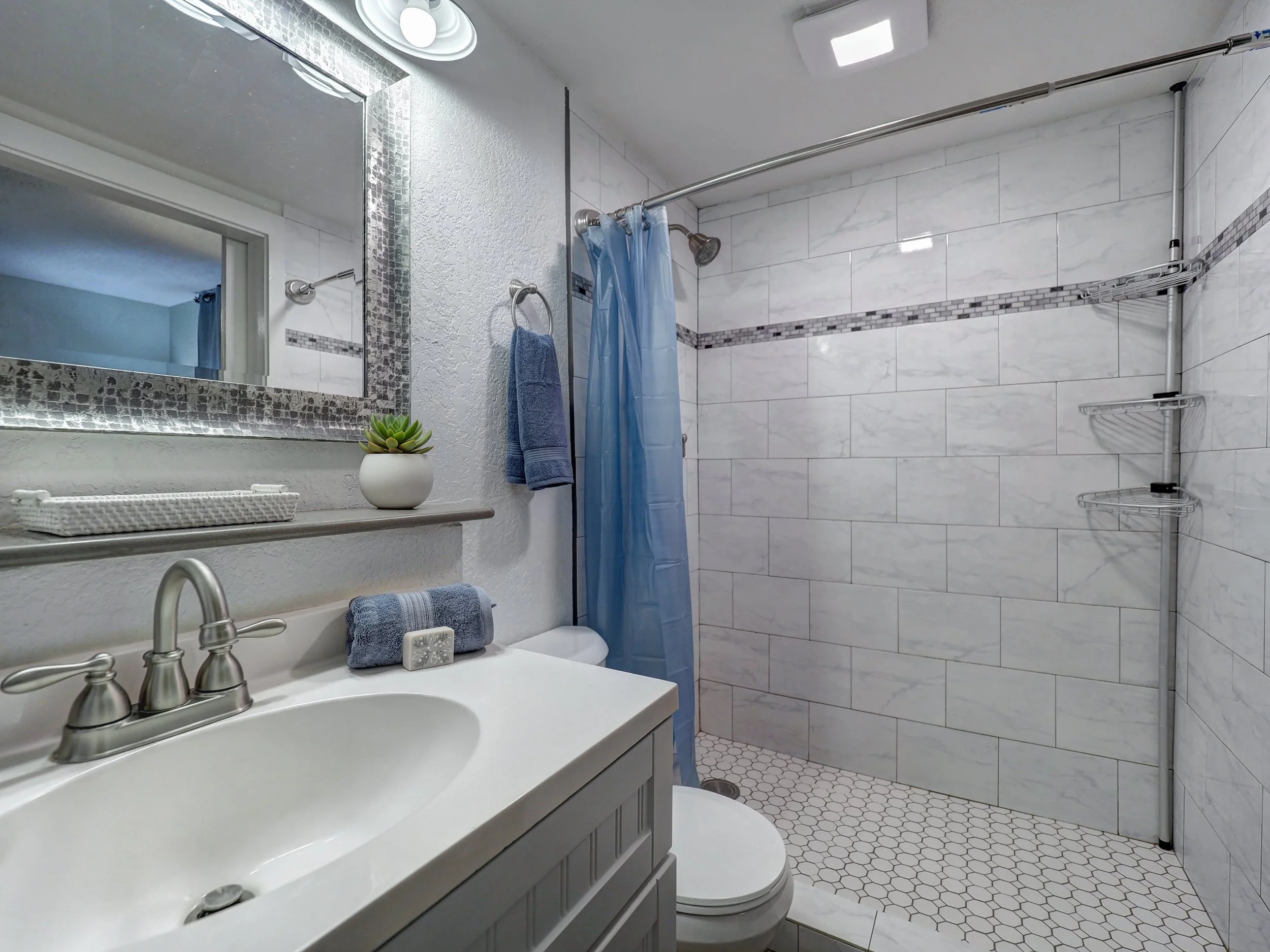 A modern bathroom with a white vanity, silver faucet, and a mirror above it. On the vanity, there is a small potted succulent plant and rolled blue towels. The bathroom features white textured walls and a shower area with a blue shower curtain. Insid