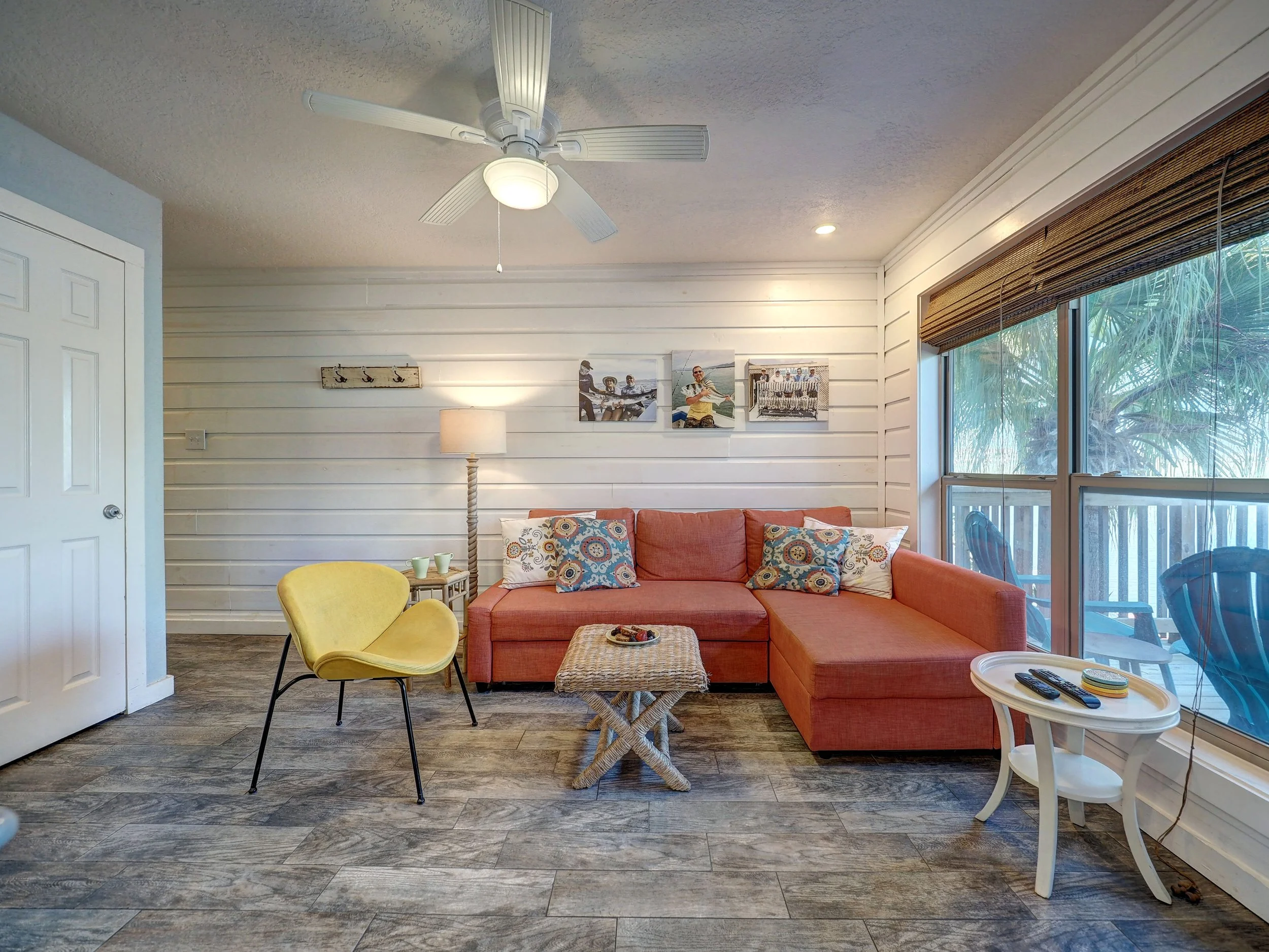 A cozy living room with white horizontal shiplap walls, a ceiling fan with a light, a sofa with colorful pillows, a yellow chair, a small side table with cups, a floor lamp, and framed photos and artwork on the walls. There is a large window with wooden blinds showing greenery outside.