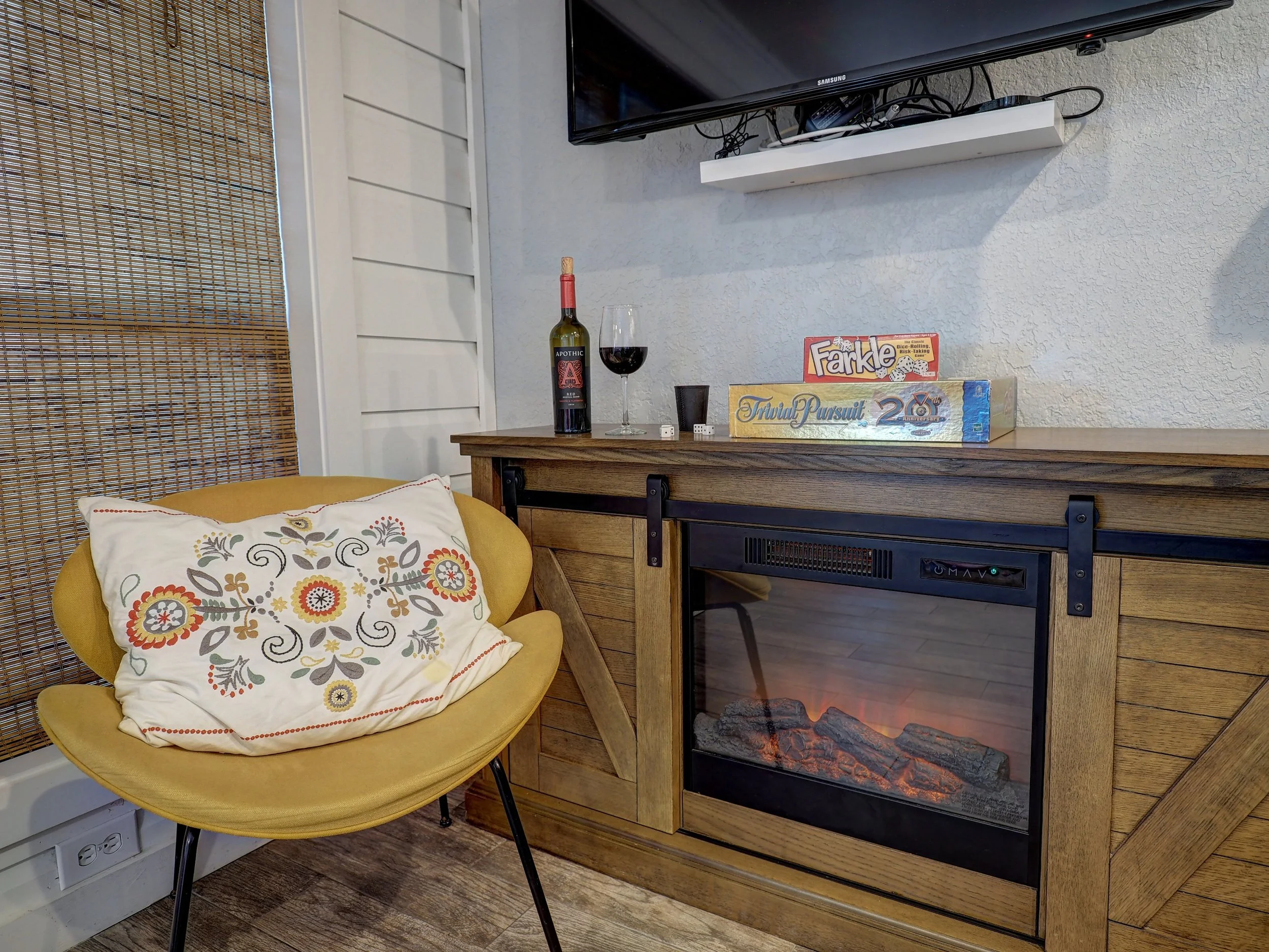 A cozy living room corner featuring a yellow chair with a colorful embroidered pillow next to a wood-paneled fireplace with a fake fire. There is a backing made of bamboo on the wall and a wall-mounted TV above the fireplace. On the wooden surface in