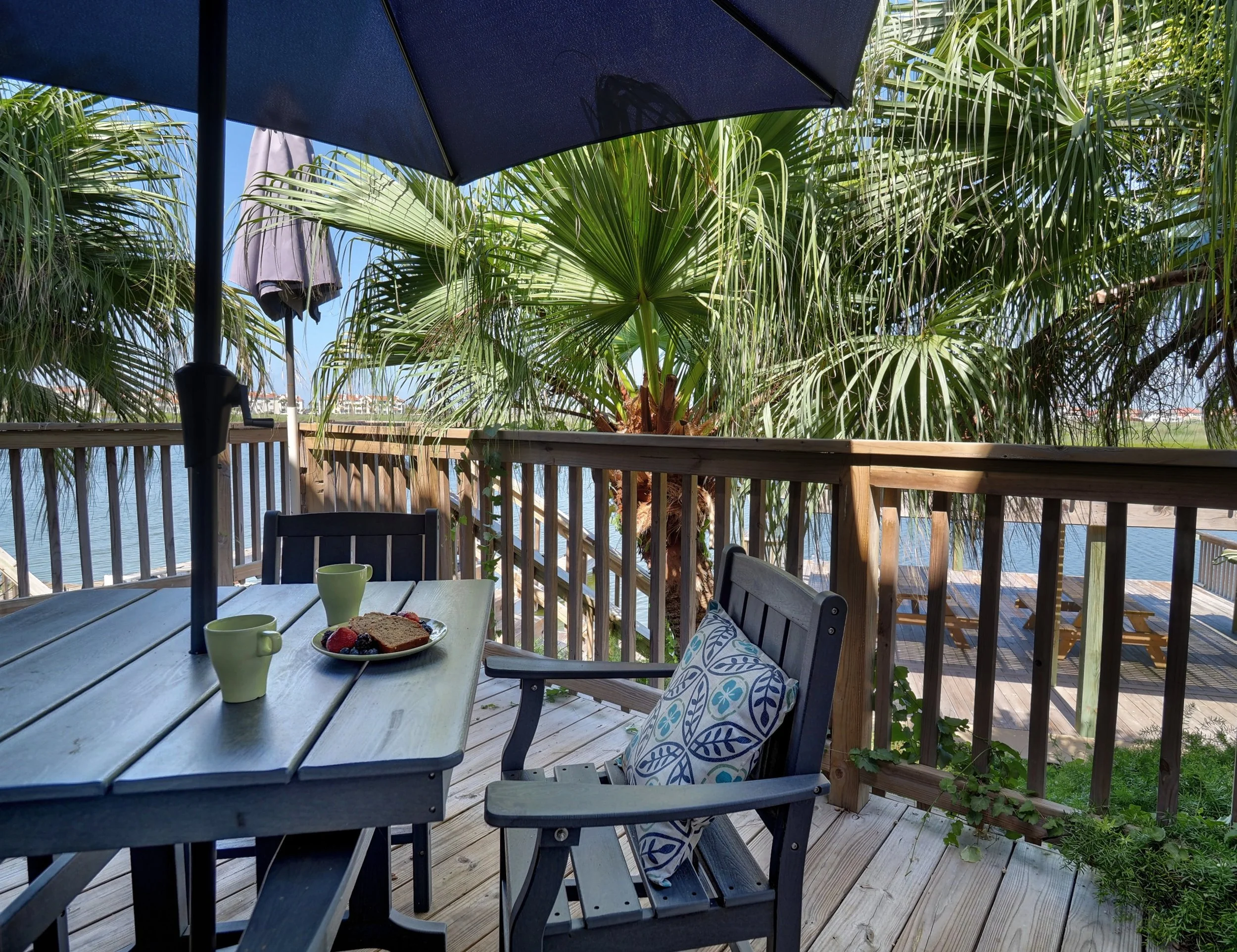 A wooden outdoor deck with a table set for breakfast with two mugs and a plate of toast with fruit, shaded by a large umbrella, overlooking lush green tropical plants near a body of water.