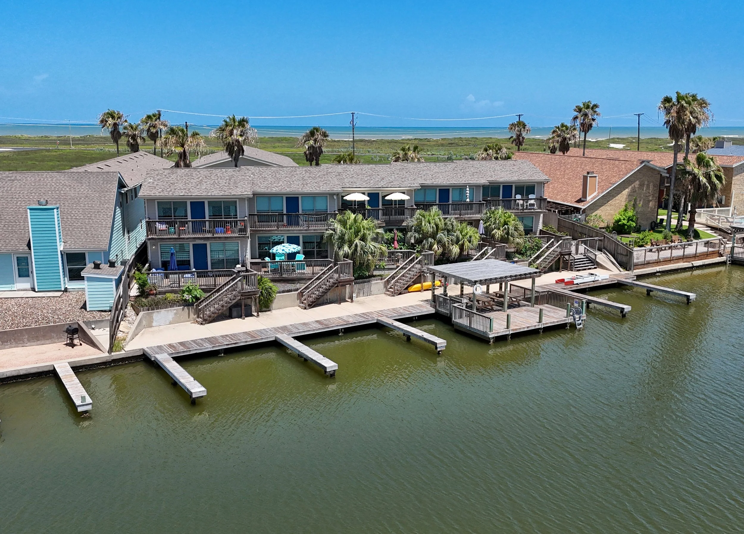 A view of rooftops of houses near a waterway with docks, and a background of open land with palm trees and a distant ocean under a blue sky.