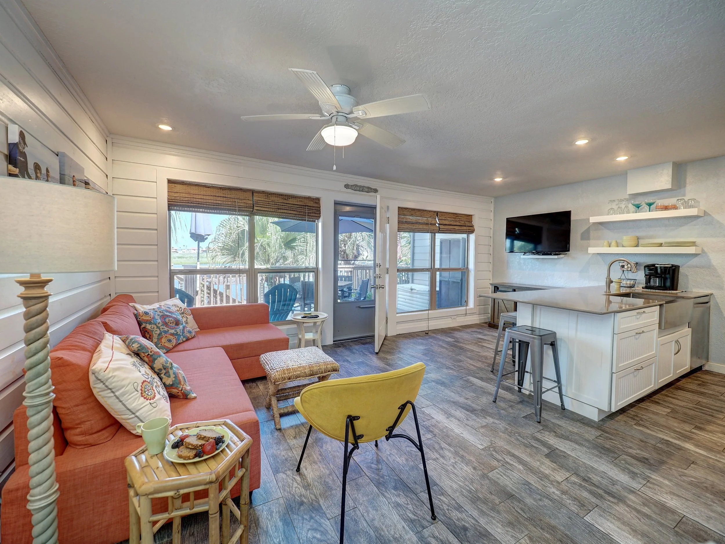 Interior of a bright, cozy living space with a peach sofa, colorful pillows, a yellow chair, a small bamboo tray with snacks, a lamp, and a kitchen area with open shelves, a TV, and a countertop.