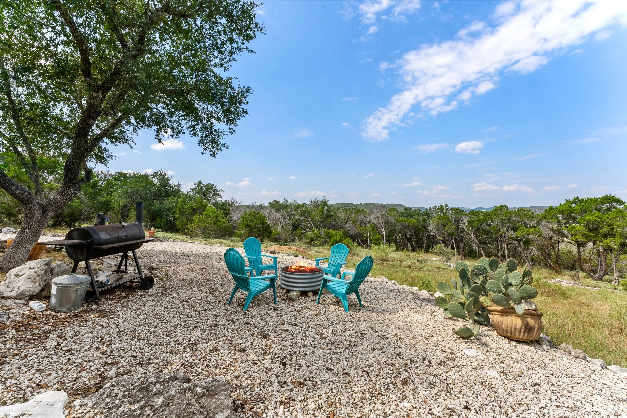 An outdoor patio area featuring a fire pit surrounded by four blue Adirondack chairs, a barbecue grill, and a cactus in a large planter, with trees and a blue sky with clouds in the background.