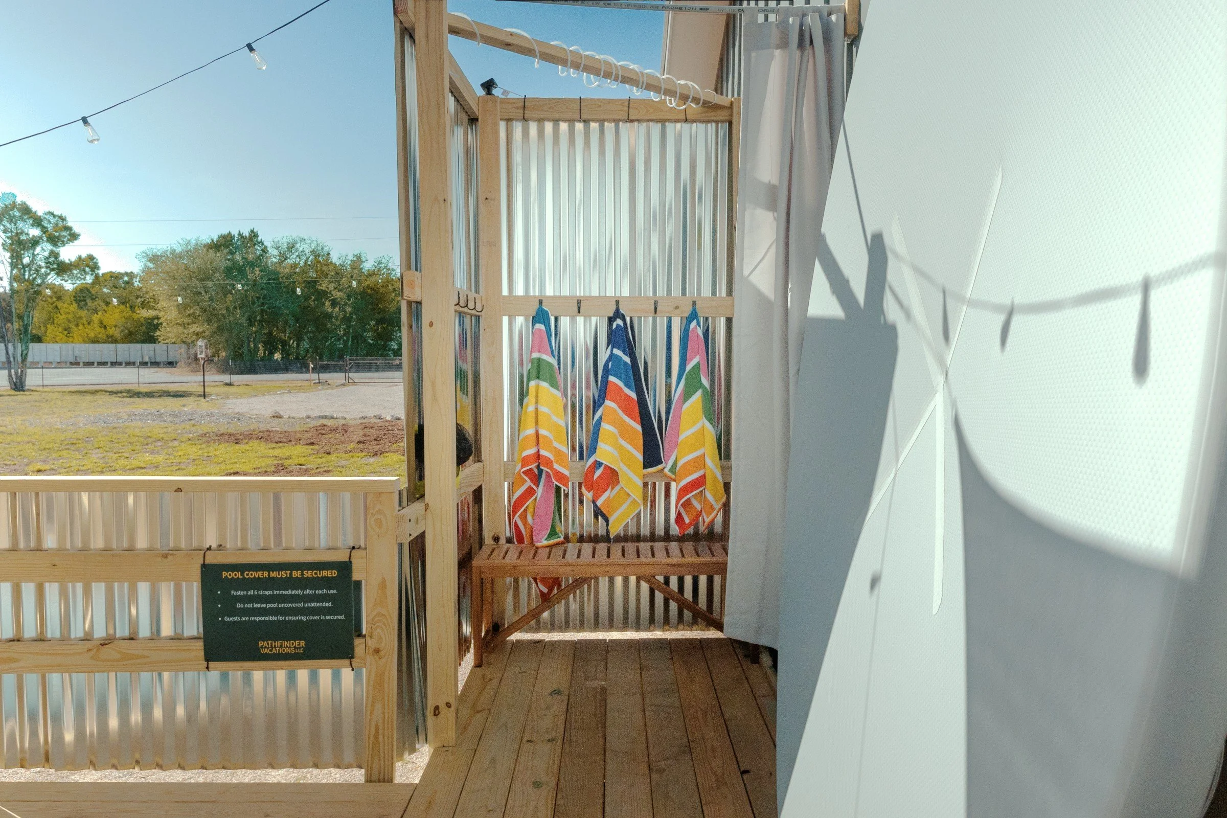 A small outdoor shower area with a wooden frame, several colorful striped towels hanging on hooks, string lights, and a corrugated metal fence, overlooking a grassy field.