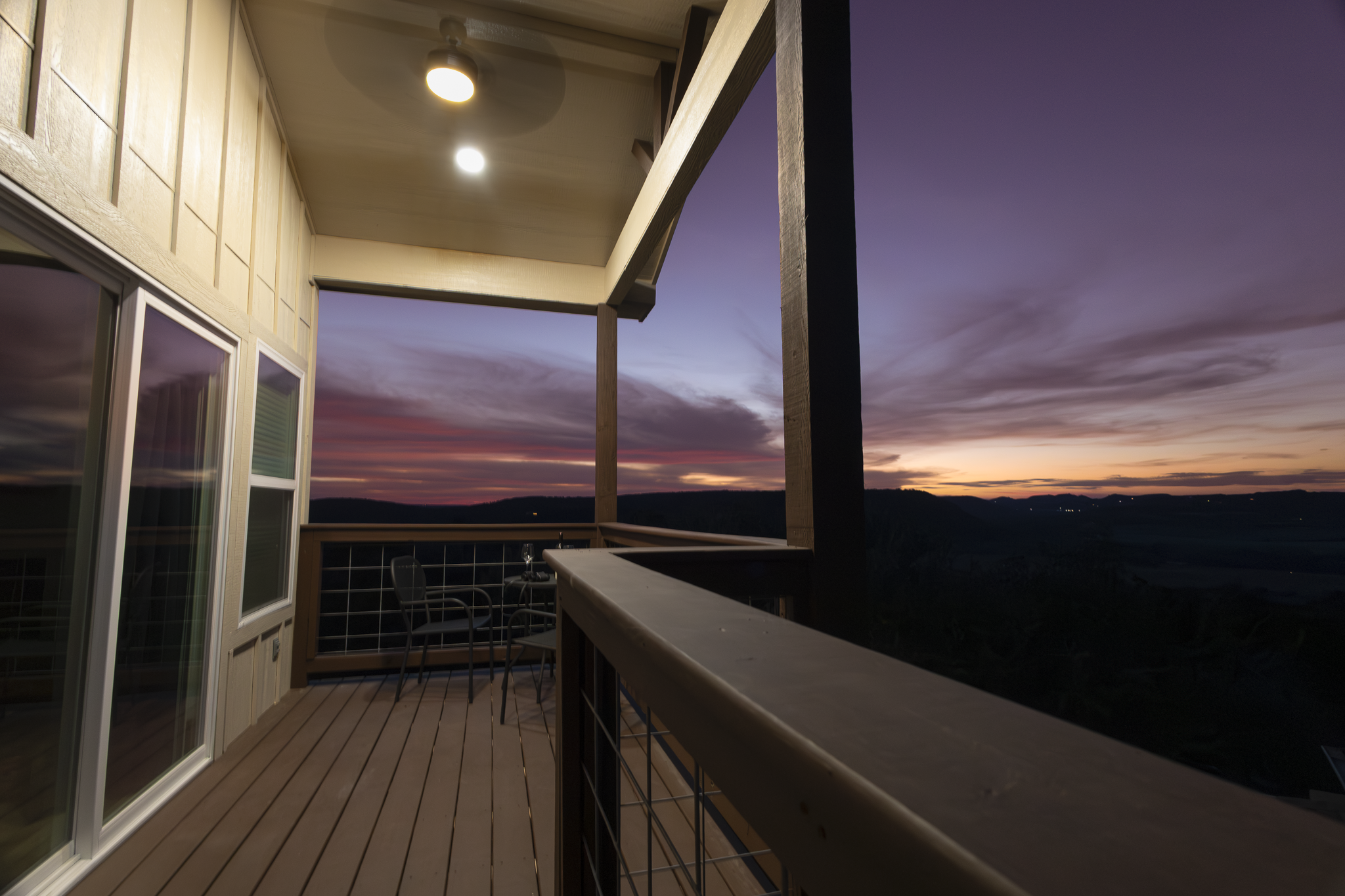 Balcony with wooden flooring and railing overlooking a sunset sky with purple and orange hues. There is a ceiling fan and recessed lighting on the ceiling, and a small seating area with two chairs and a table.
