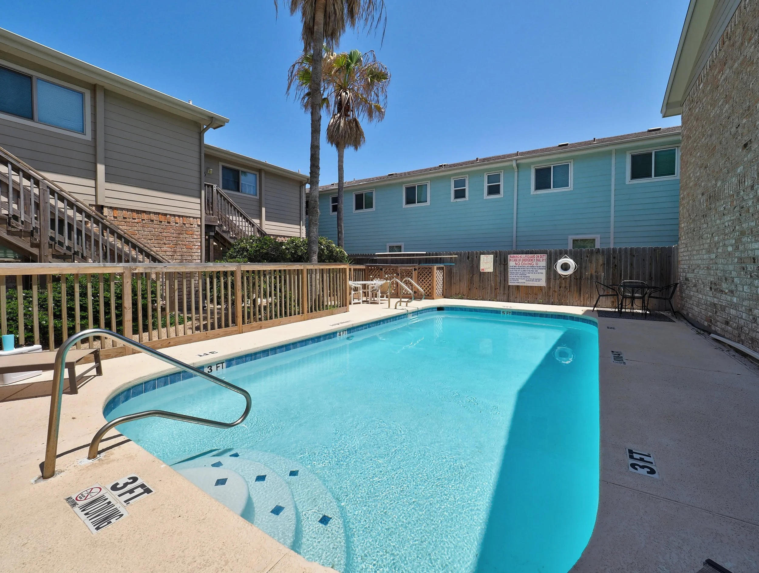 A swimming pool with a wooden fence, blue water, and a concrete deck. Two tall palm trees are visible, with one showing signs of dying fronds. Surrounding buildings with beige and light blue siding and brick walls are in the background. There are pla