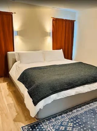 A neatly made bed with white sheets, a dark gray blanket, and white pillows, in a room with beige walls, wooden curtains, and wall-mounted lights.