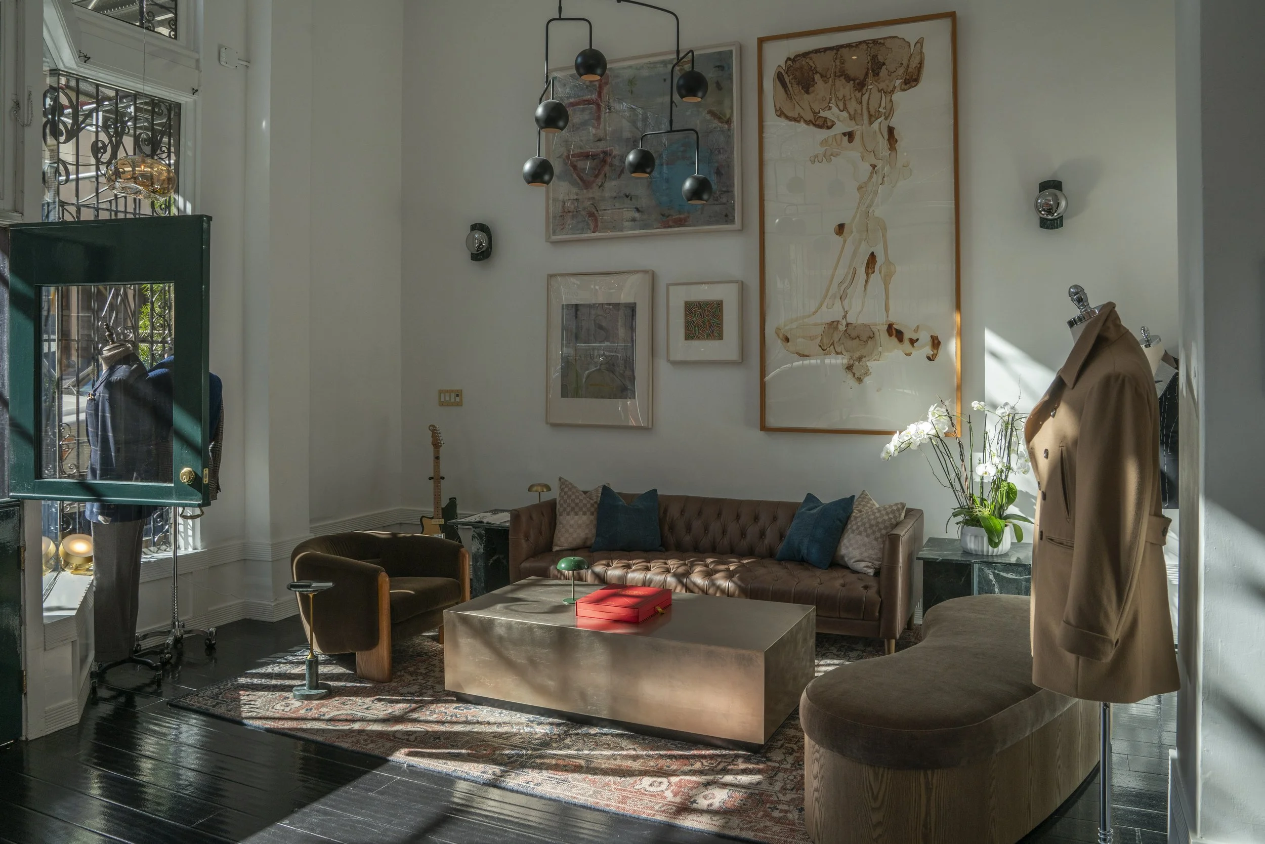 A stylish lounge in a showroom with a brown leather tufted sofa, a coffee table with a red box, a potted orchid, artwork on the wall, a mannequin with a brown blazer, and a window with sunlight streaming in.