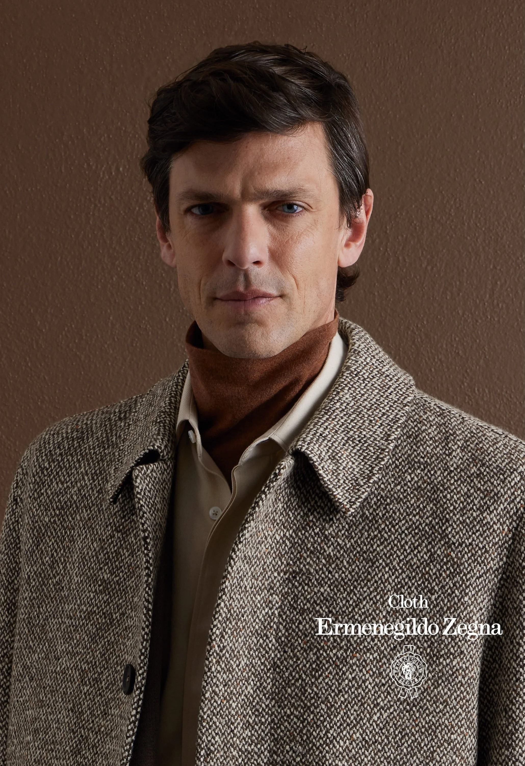 Man wearing a brown checkered coat and a beige shirt with a brown turtleneck underneath, against a brown textured wall, with white text on the image that reads 'Cloth Ermenegildo Zegna' and a circular emblem.