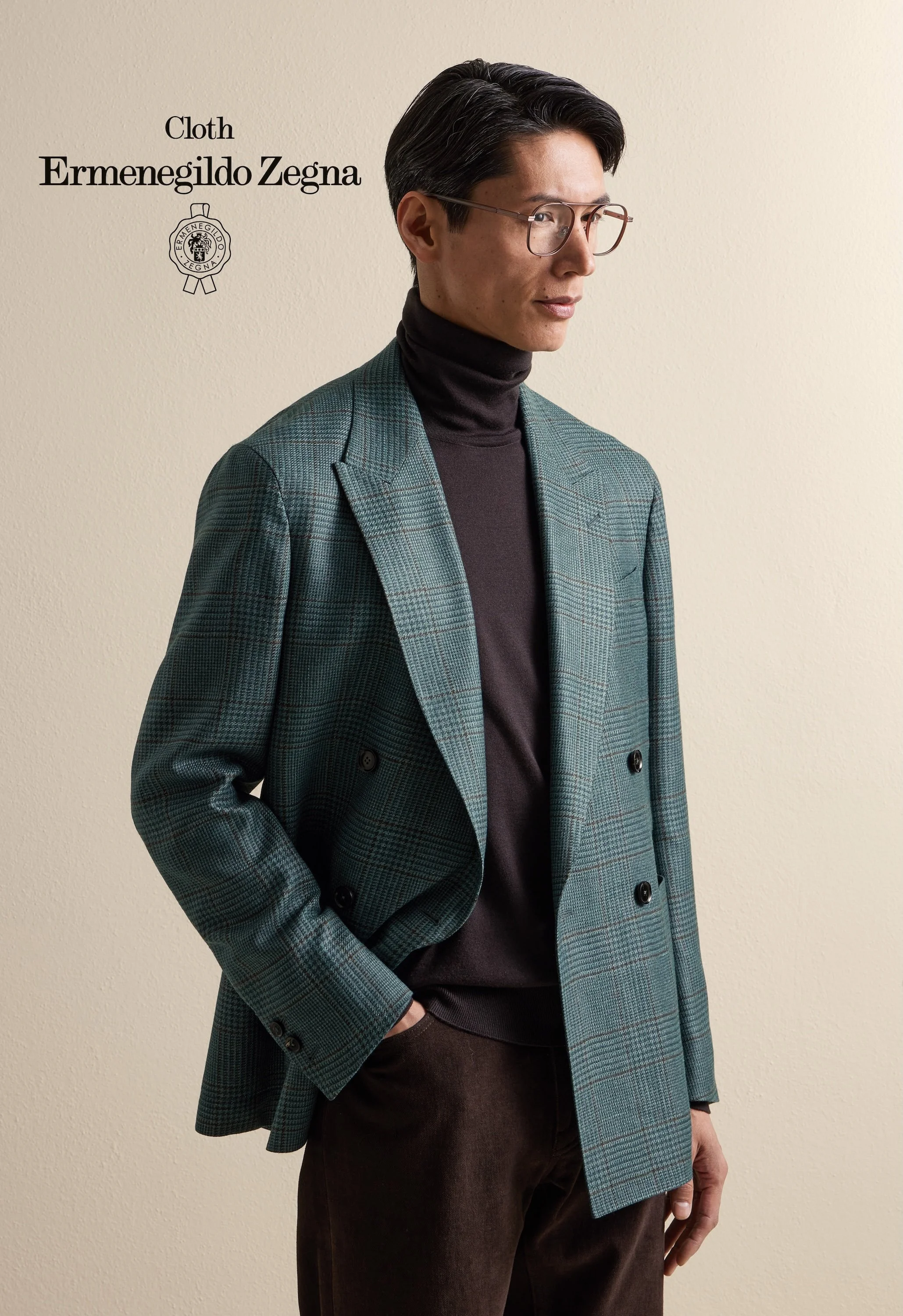 A man wearing glasses, a black turtleneck, a green plaid Zegna blazer, and brown pants stands against a beige wall.