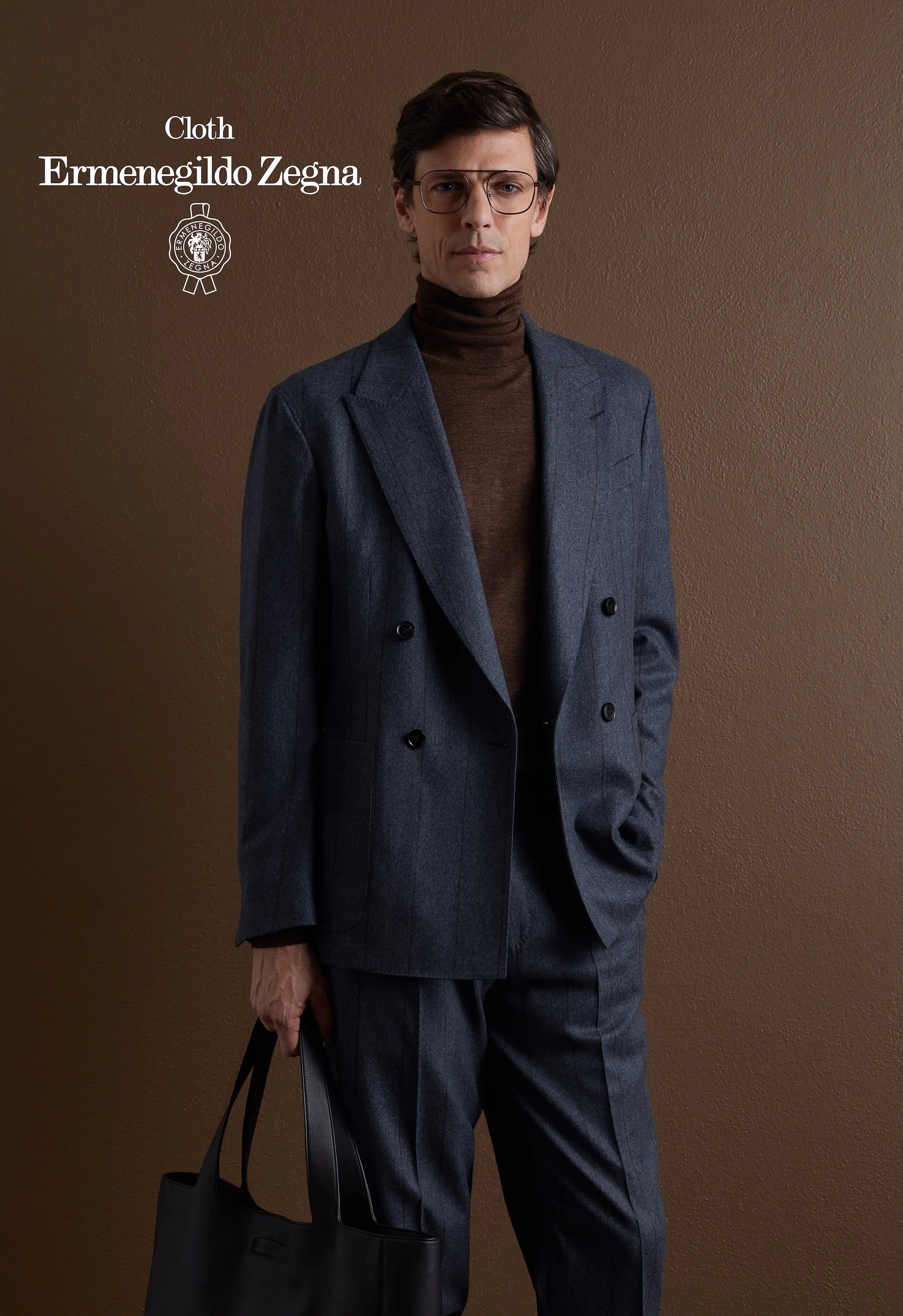 A man in a dark gray plaid Zegna suit, brown turtleneck, and glasses holding a black leather bag, standing against a brown wall.