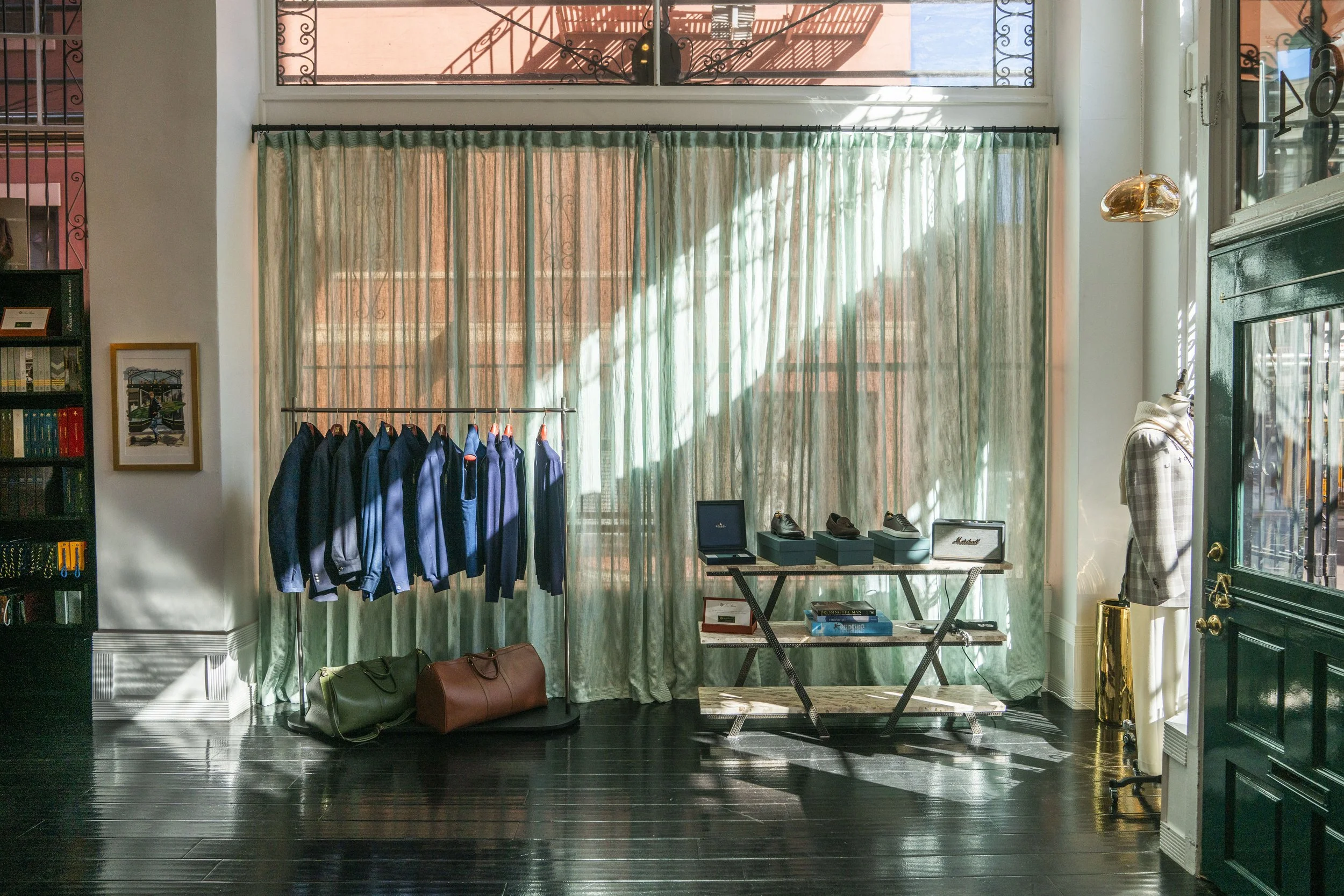 Interior view of the showroom with a rack of jackets, a window with green curtains, a table displaying shoes, and various bags on the floor. Sunlight streams through the window, casting shadows on the dark wooden floor.