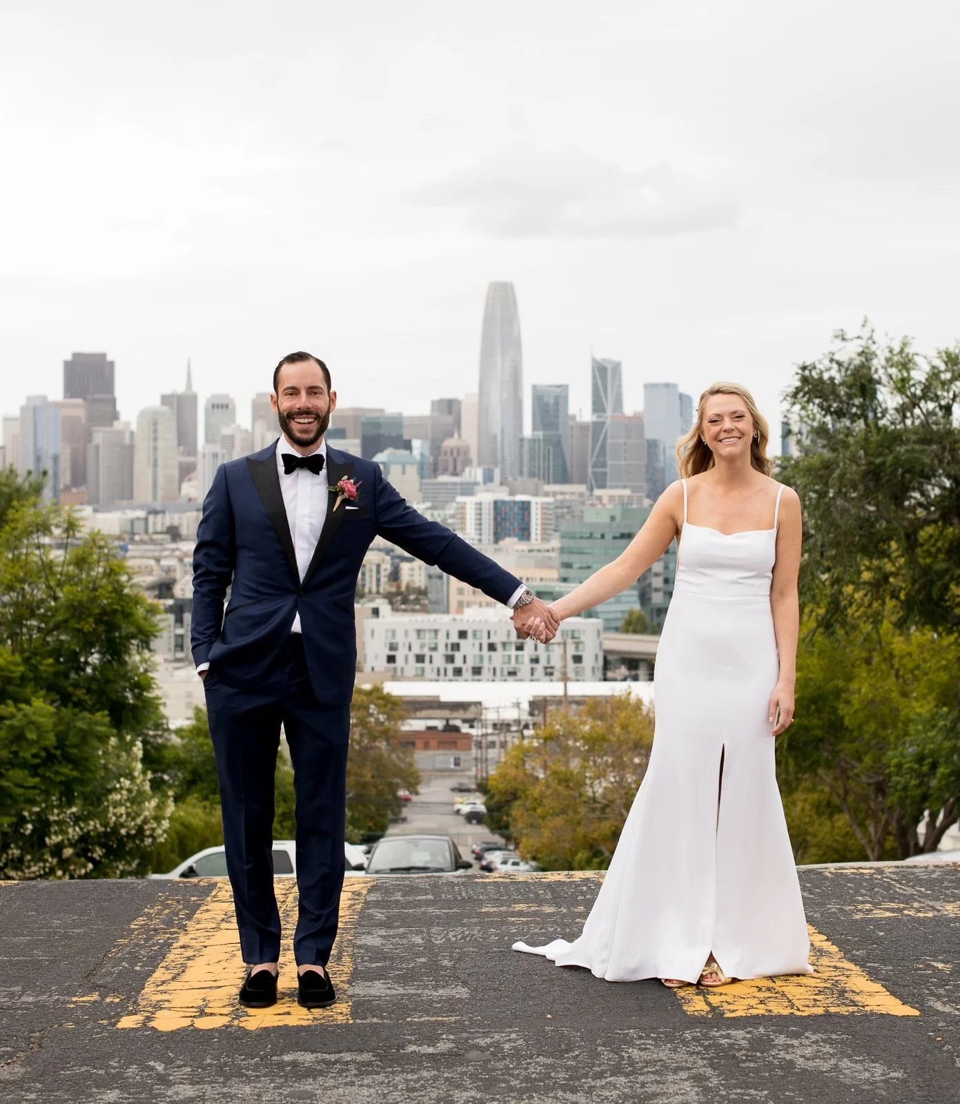 A newlywed couple smiling and holding hands in a city parking lot with a city skyline in the background, including tall buildings and skyscrapers, on an overcast day with the groom wearing a Scarborough custom suit.