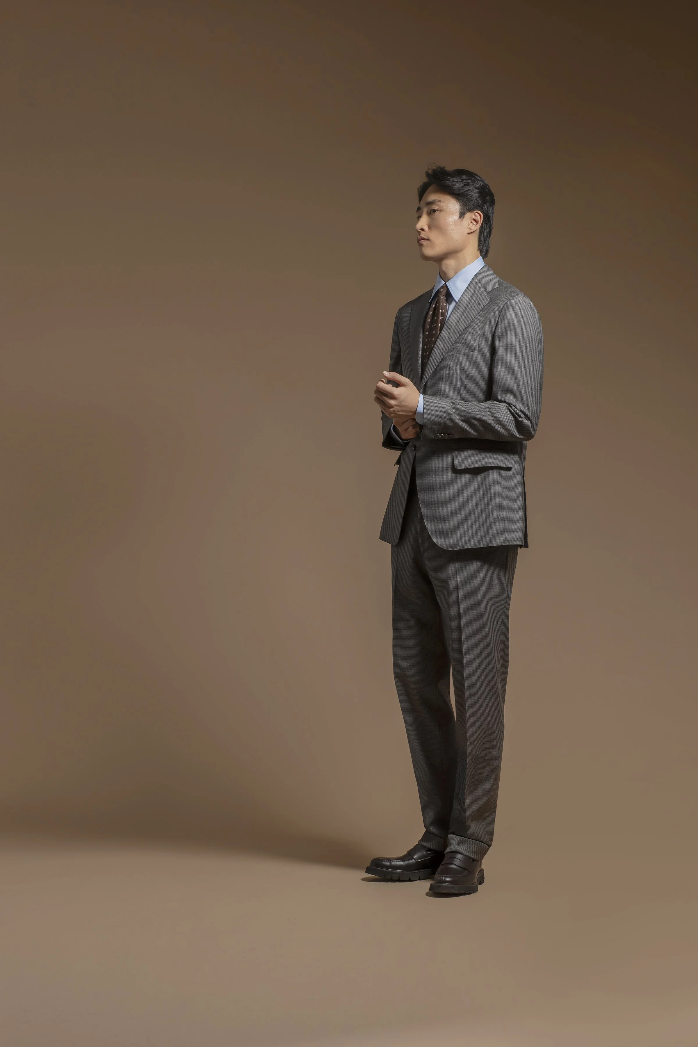 A man in a tailored Scabal gray plaid suit with a light blue shirt, dark brown polka dot tie, and black leather shoes, standing against a plain brown background, looking to the side with a serious expression.