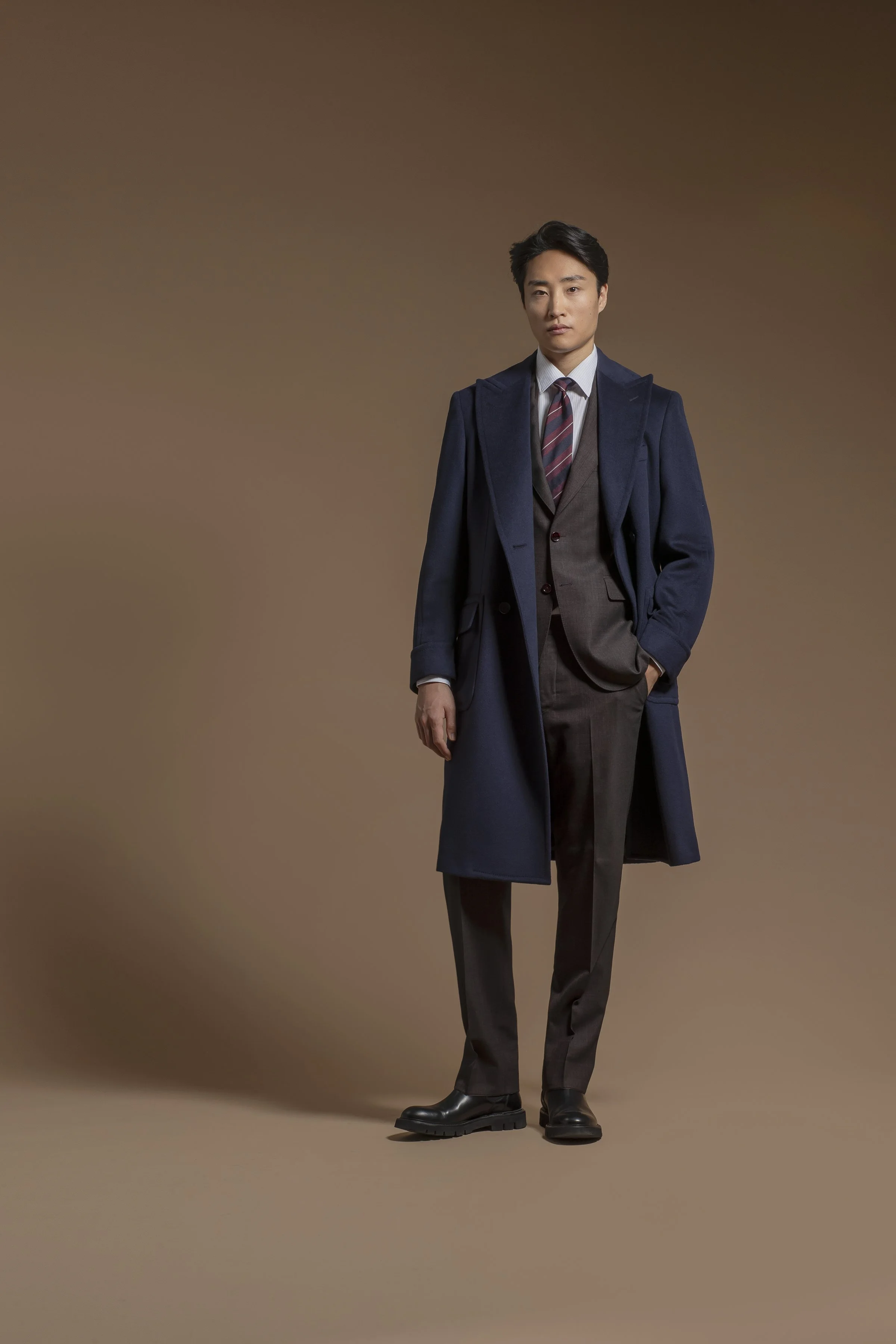 A young man in Scabal formal attire, including a blue overcoat, gray suit, tie, and dress shoes, standing against a plain brown background.