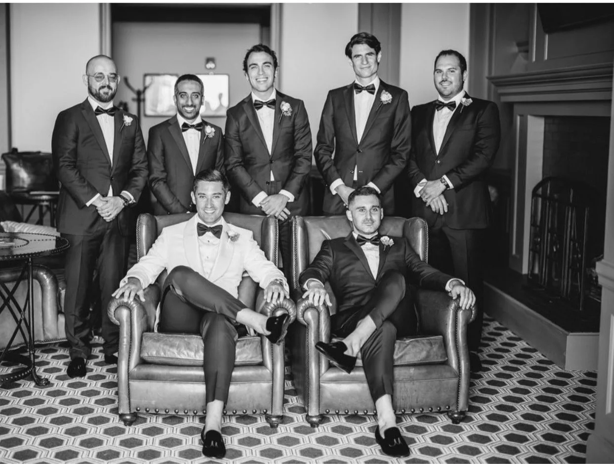 Black and white photo of a groom and wedding party in Scarborough Custom Tailoring suits and tuxedos, some with boutonnières, in a room with a fireplace, patterned carpet, and window. Two men seated in front on a sofa, while five men stand behind.