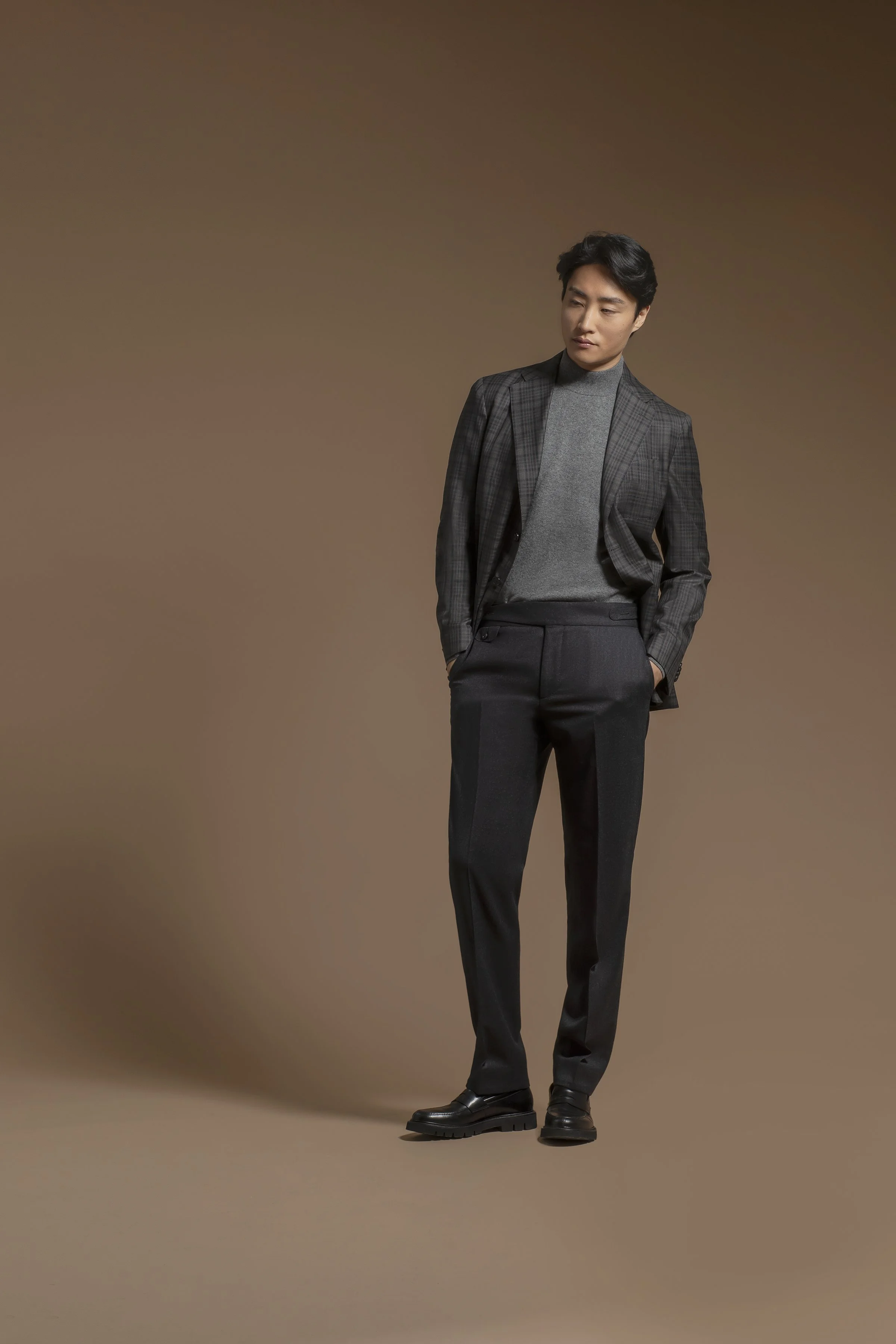 A man wearing a Scabal dark checkered blazer, gray turtleneck, and black dress pants, standing against a plain brown background.