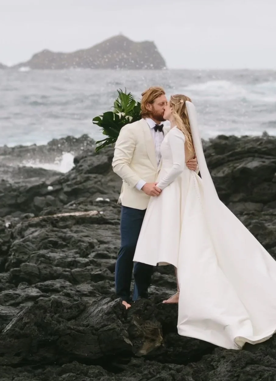 A bride and groom wearing Scarborough Custom Tailoring tuxedo share a kiss on a rocky shoreline, with an island in the background during a cloudy day.