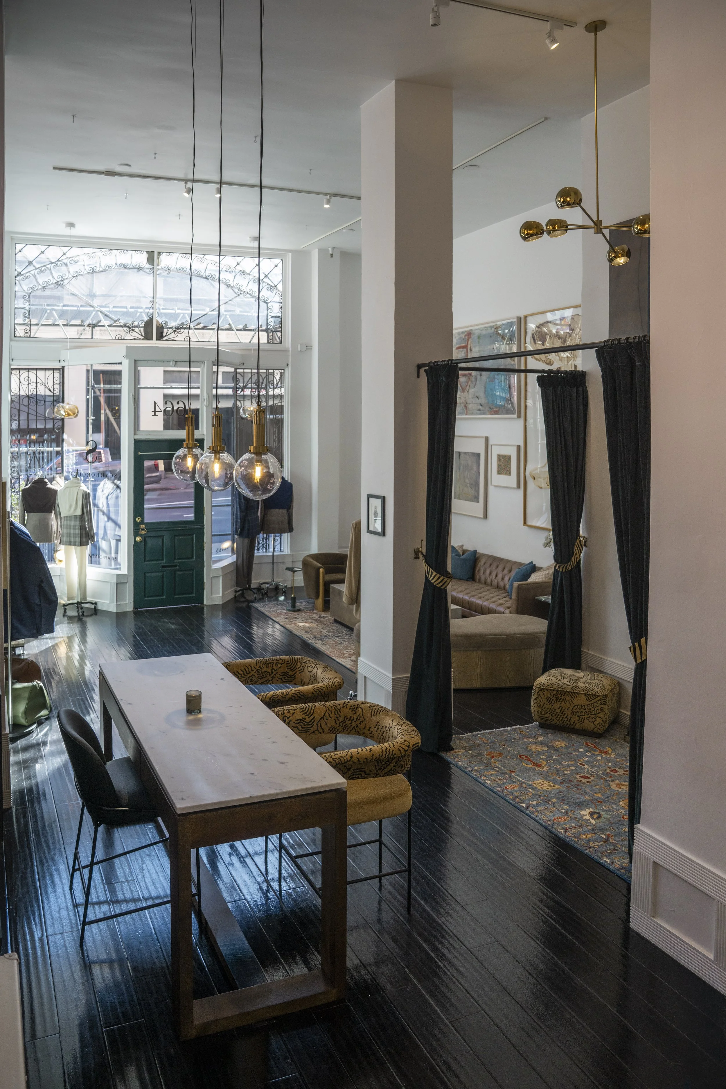 Interior of the showroom with a high ceiling, large front windows, and various furniture including a marble table with chairs, a vintage sofa, framed artwork on the walls, and stylish lighting fixtures.
