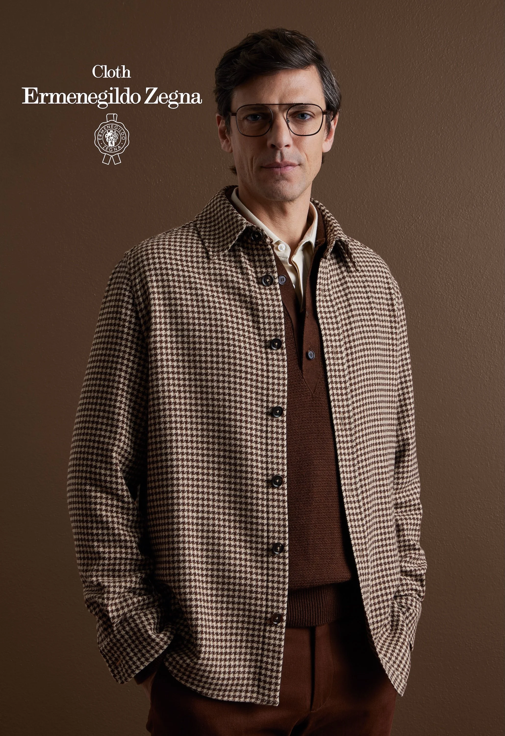A man with dark hair wearing glasses, a beige and brown checkered Zegna jacket, and a brown sweater standing against a plain brown wall.